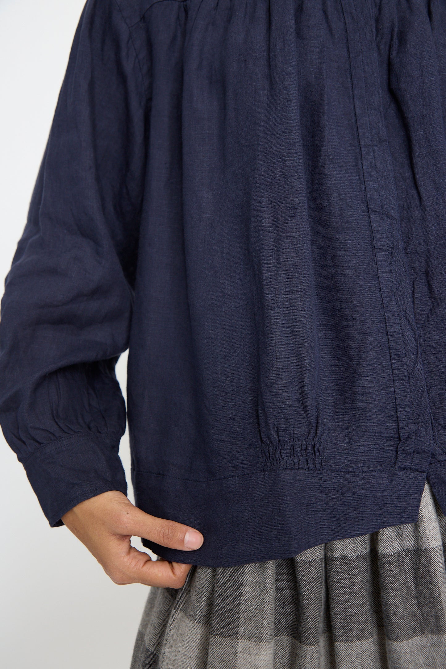 A close up of a person wearing the Linen Shirt in Navy by Ichi Antiquités with a gray plaid skirt, holding the hem of the blouse with one hand.