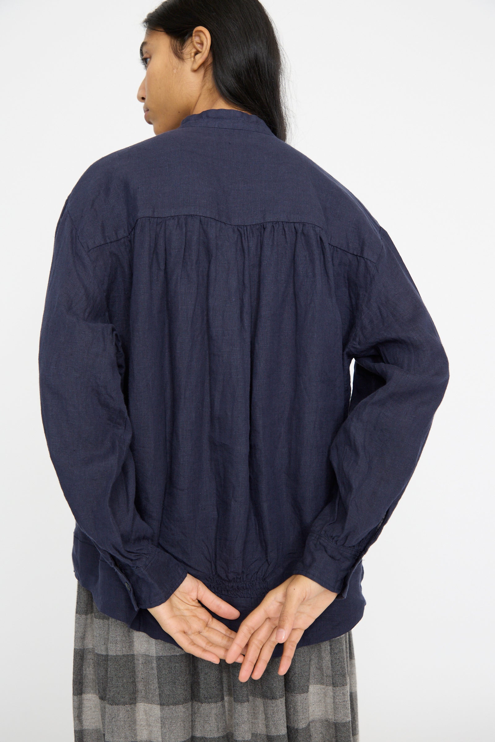 A person in an Ichi Antiquités Linen Shirt in Navy, featuring a relaxed fit and band collar, stands with hands behind their back, facing away from the camera. They're paired with a gray checkered skirt.