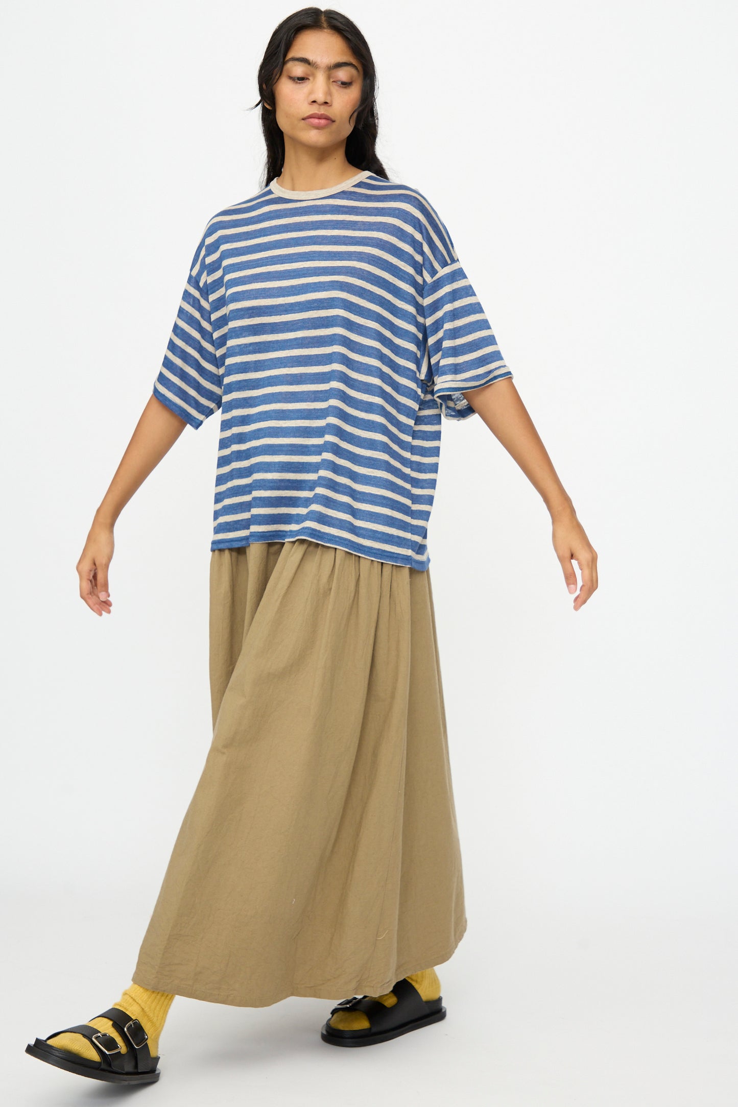 A person wears the Ichi Antiquités Linen Stripe Pullover in Blue and Natural, paired with a long tan skirt, yellow socks, and black sandals, standing against a plain white background.