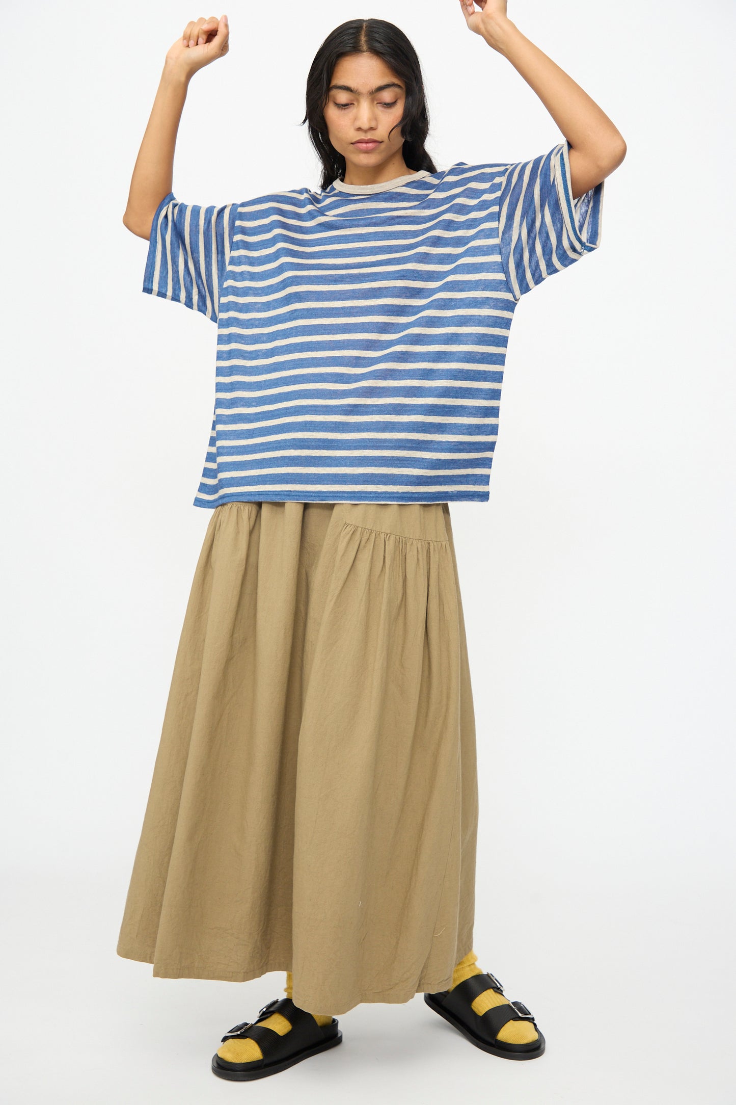 A person stands with arms raised, wearing the Ichi Antiquités Linen Stripe Pullover in Blue and Natural, paired with a long beige skirt, yellow socks, and black sandals against a plain white background.