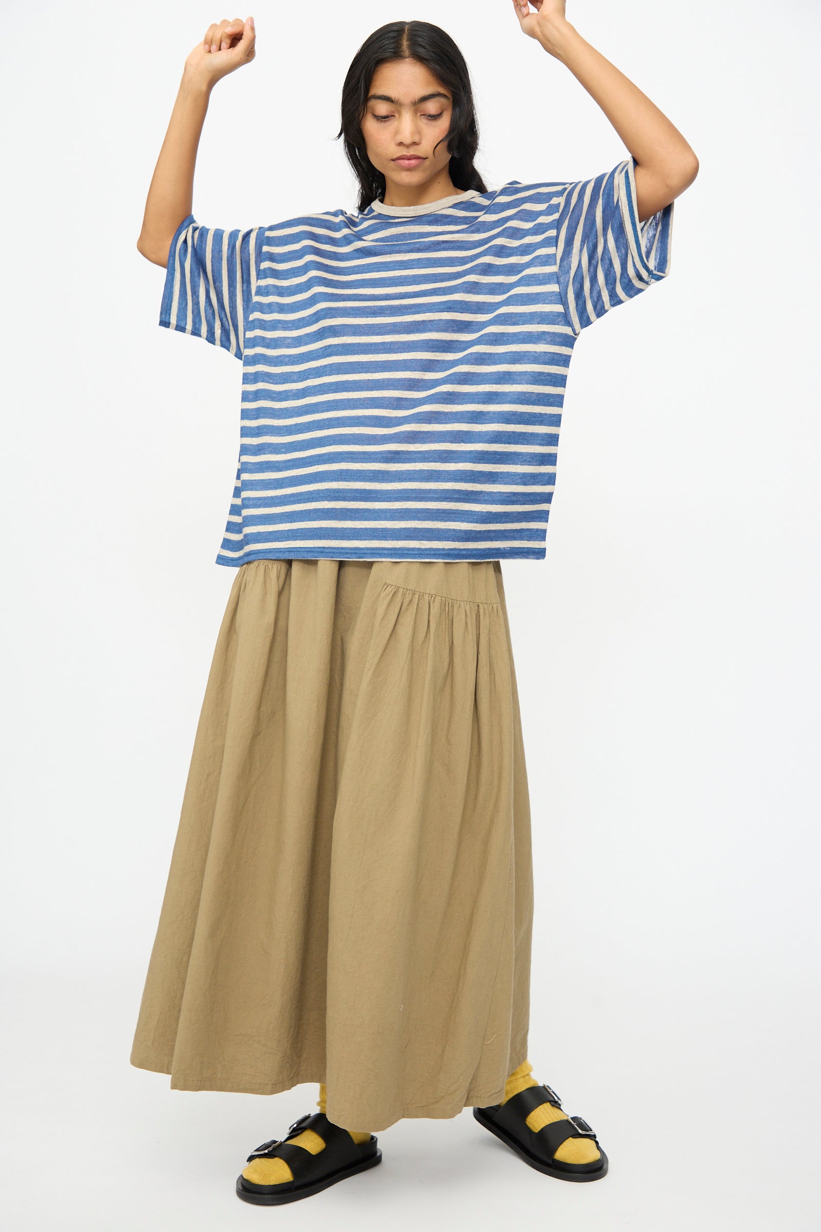 A person stands with arms raised, wearing the Ichi Antiquités Linen Stripe Pullover in Blue and Natural, paired with a long beige skirt, yellow socks, and black sandals against a plain white background.
