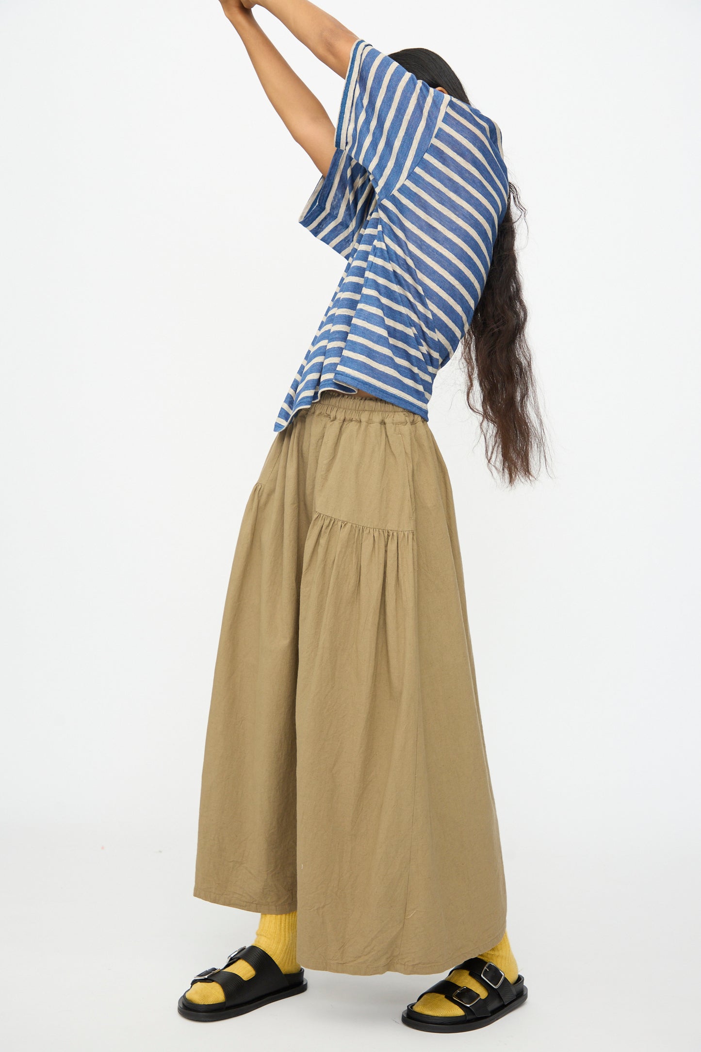 A person wearing the Ichi Antiquités Linen Stripe Pullover in Blue and Natural stands with arms raised, facing sideways against a plain white background. They pair it with a loose tan skirt, yellow socks, and black sandals.