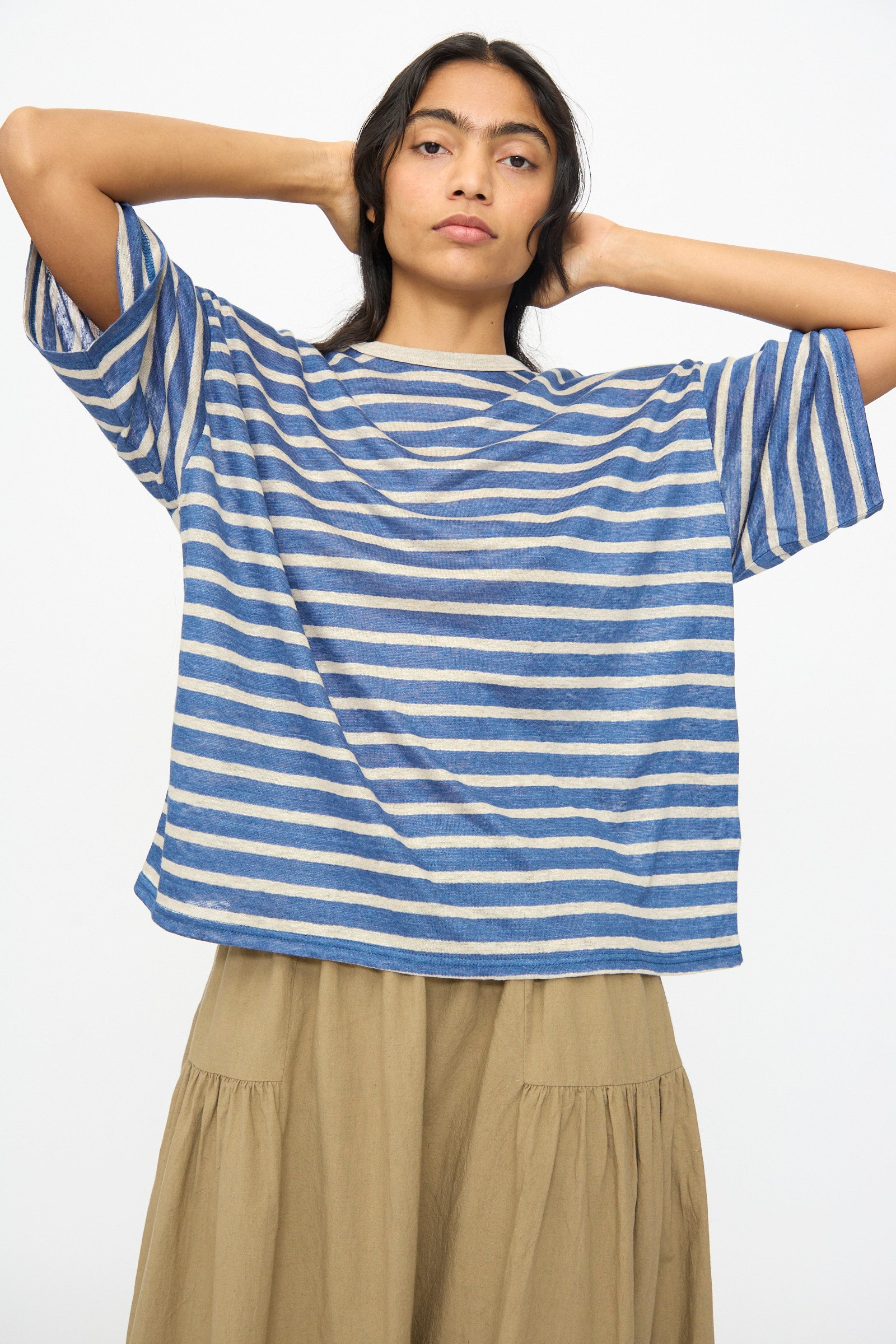 A person stands with hands behind their head, wearing the Ichi Antiquités Linen Stripe Pullover in Blue and Natural and a beige skirt, against a plain light background.