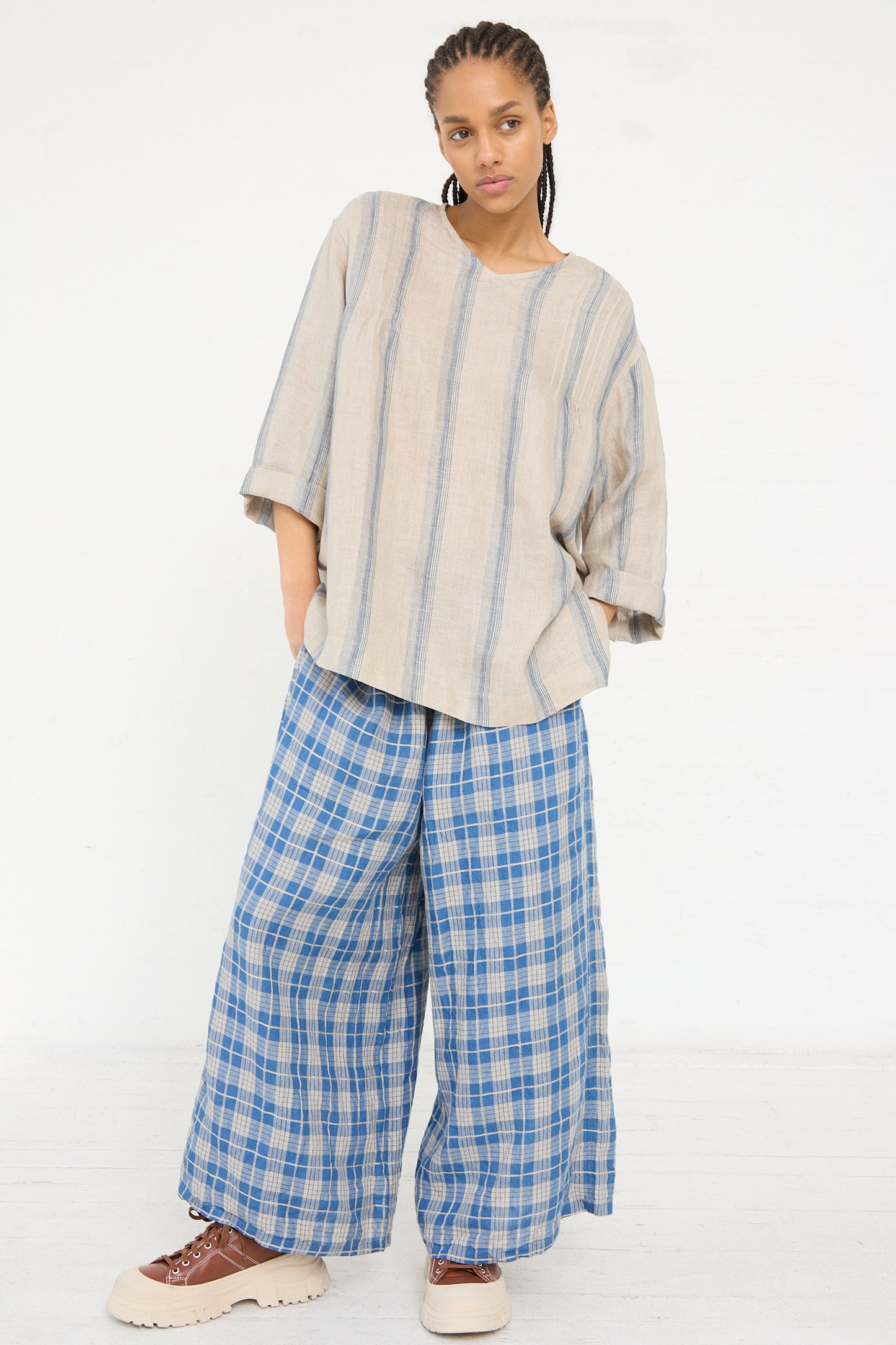 A person standing in a neutral pose wearing an Ichi Antiquités Linen Stripe Pullover in Natural and Indigo with wide blue checkered trousers and lace-up shoes.