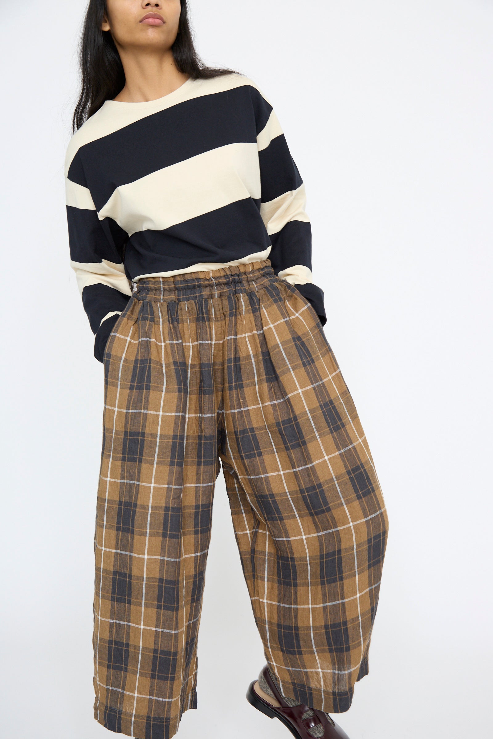 A person stands against a plain background wearing a black and white striped long-sleeve shirt paired with Ichi Antiquités' Linen Tartan Pant in Brown, featuring a wide-leg style with a subtle plaid check pattern.