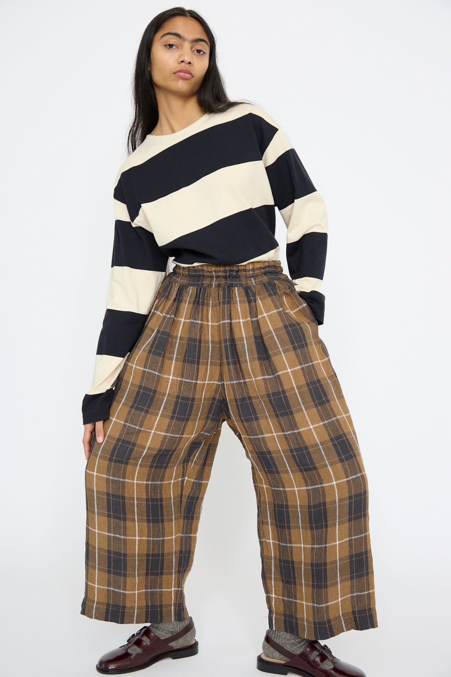 A person wears a black and white striped long-sleeve shirt with Ichi Antiquités Linen Tartan Pant in Brown, standing on a white background with legs standing wide. 