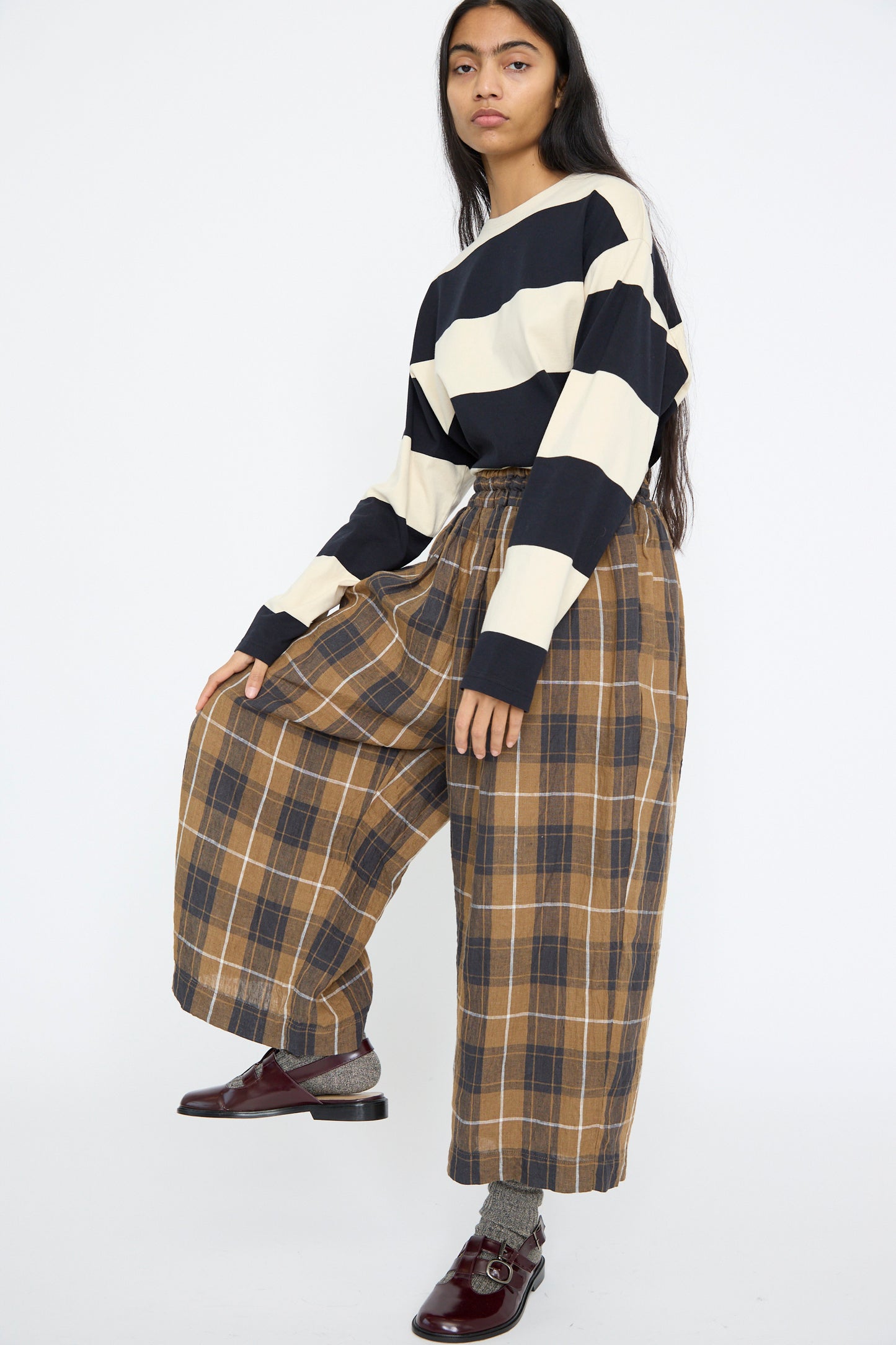 A person in a striped long-sleeve shirt and Ichi Antiquités Linen Tartan Pant in Brown poses with one leg raised, against a white background. 