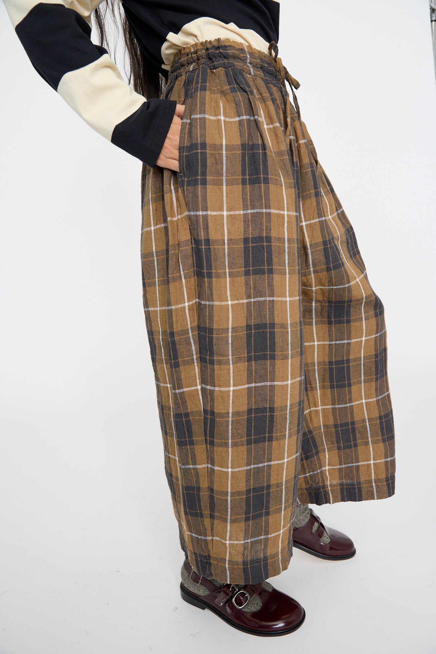 A model wearing Ichi Antiquités' Linen Tartan Pant in Brown with a shirred waist, a striped top, and shiny burgundy shoes.