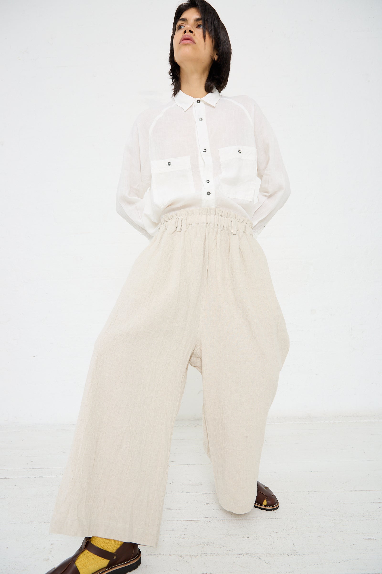 Person standing against a white background, wearing a relaxed fit white button-up shirt and Linen Vintage Pant in Natural by Ichi Antiquités, paired with yellow socks and brown sandals.