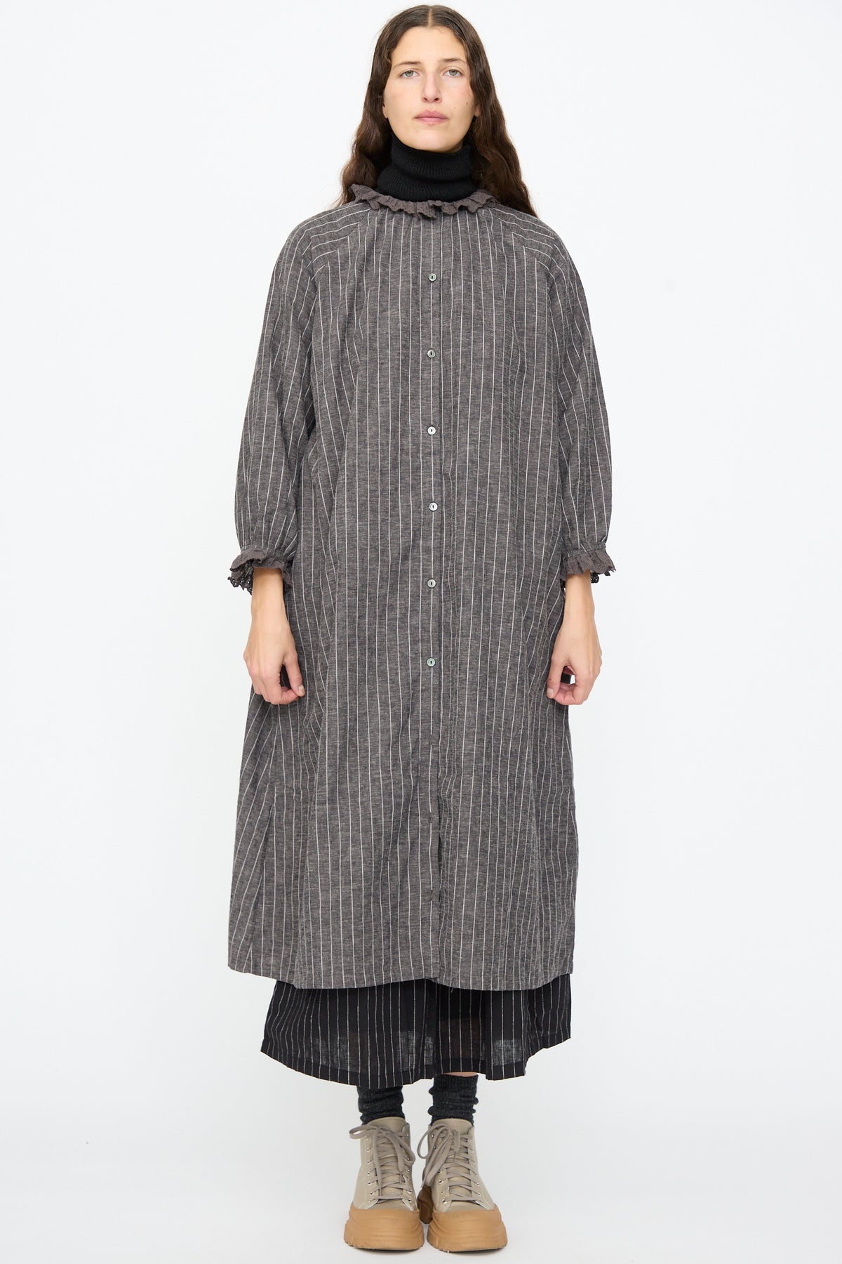A person wearing an Ichi Antiquités Pinstripe Dress in Charcoal with a lace ruffle collar and cuffs over a black pinstripe skirt and beige boots.