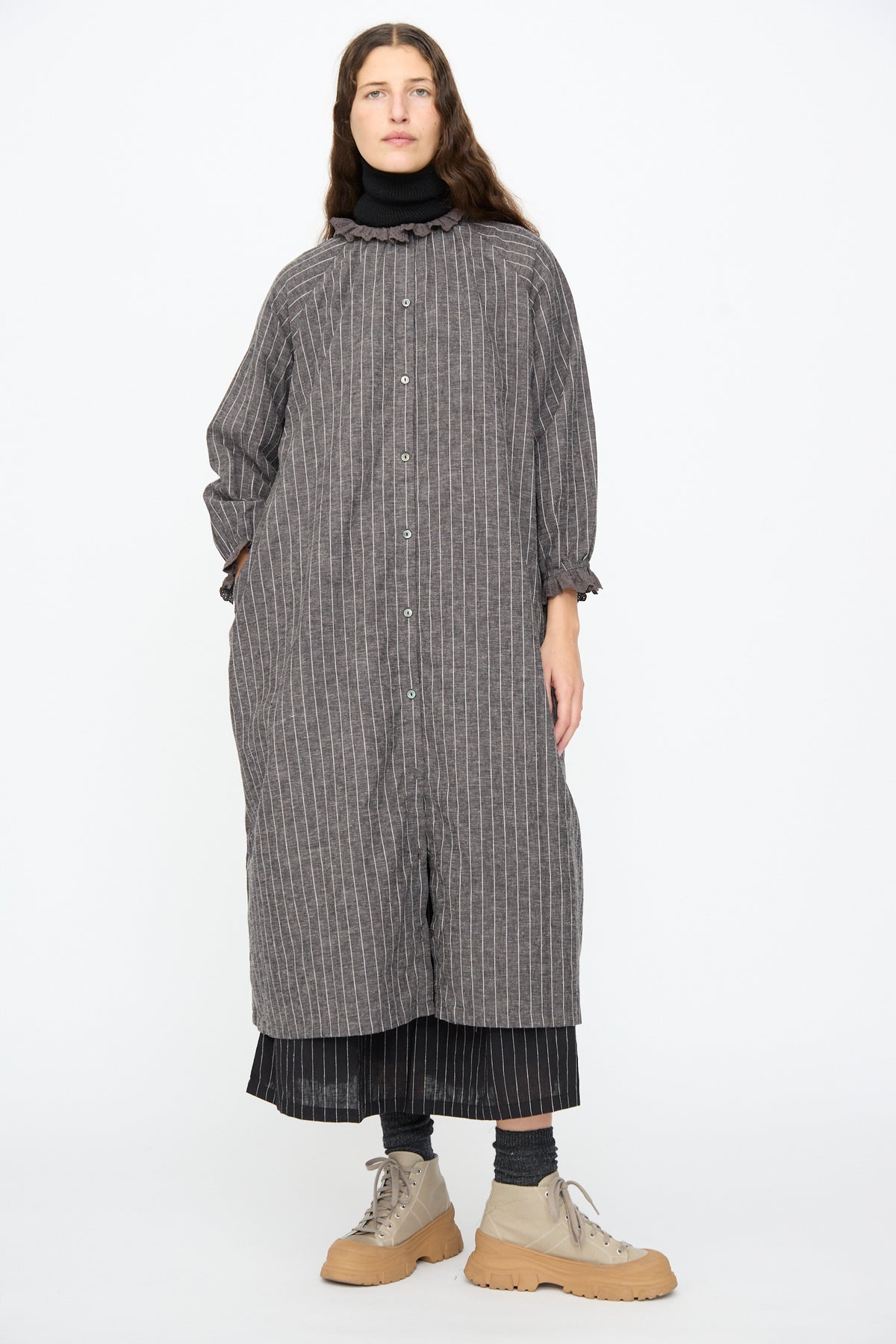 A person wears a charcoal grey pinstripe linen Pinstripe Dress in Charcoal by Ichi Antiquités with a lace ruffle collar, grey socks, and beige lug-sole boots.