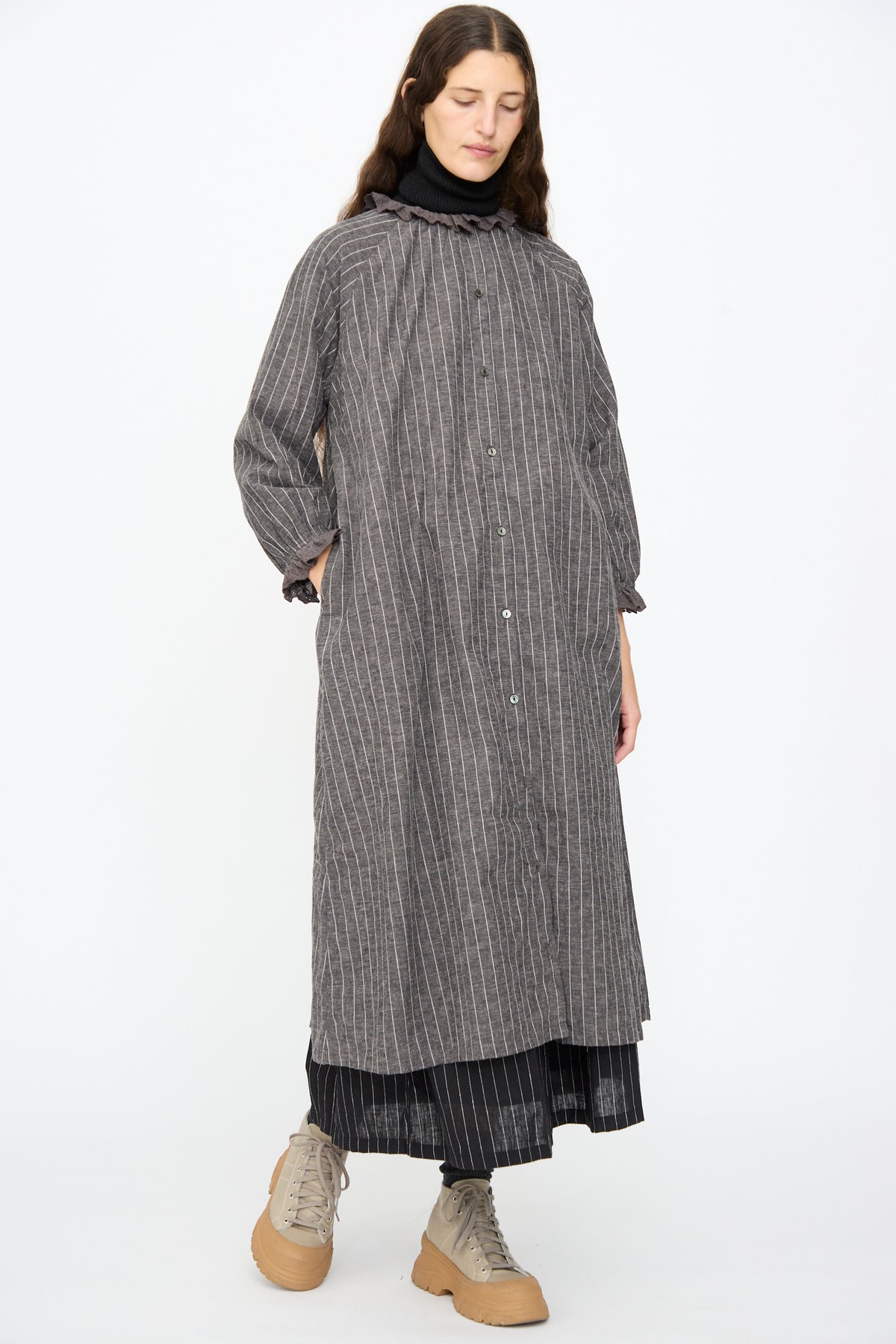A person wears an Ichi Antiquités Pinstripe Dress in Charcoal over a black turtleneck and skirt, finished with beige lace-up boots.