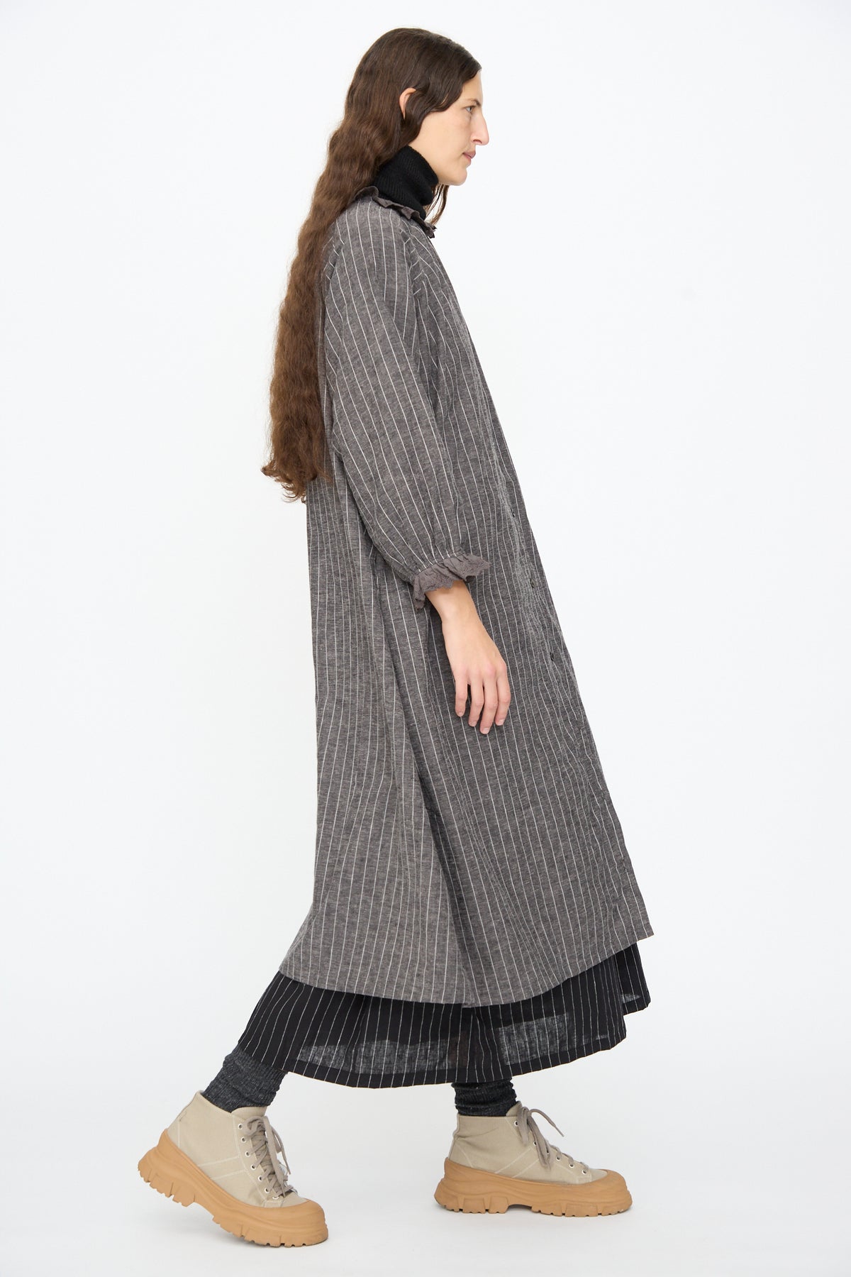 A person wearing an Ichi Antiquités Pinstripe Dress in Charcoal over a dark pleated skirt, a black turtleneck, and beige lace-up boots.