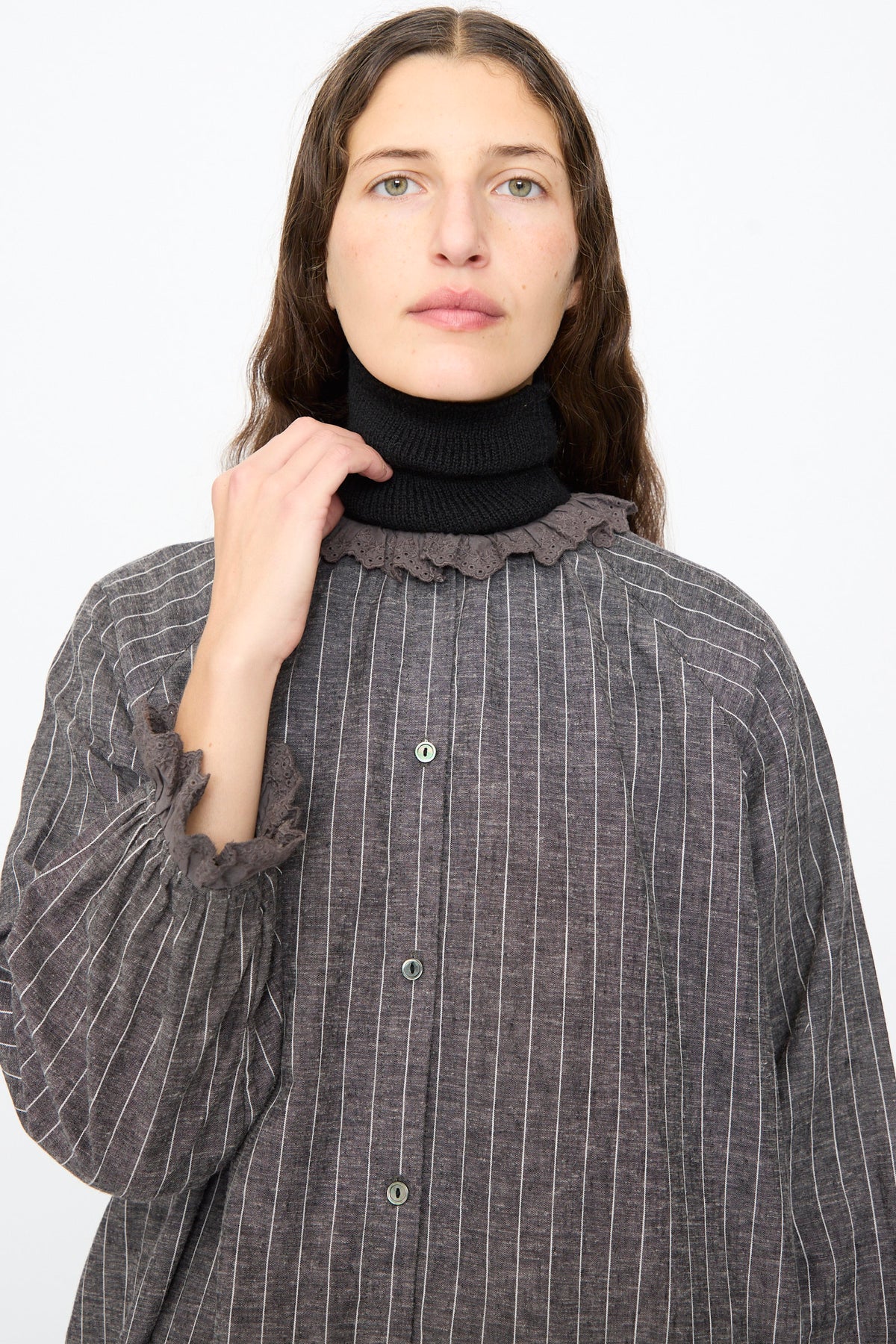 A person wears an Ichi Antiquités Pinstripe Dress in Charcoal, a charcoal grey pinstripe linen dress with a lace ruffle collar and cuffs, layered over a black turtleneck.