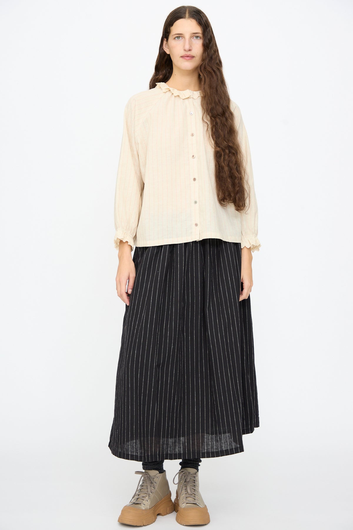 A model wears an Ichi Antiquités Pinstripe Shirt in Natural with a lace ruffle collar and cuffs, paired with a black, ankle-length pinstripe skirt and beige lace-up boots.
