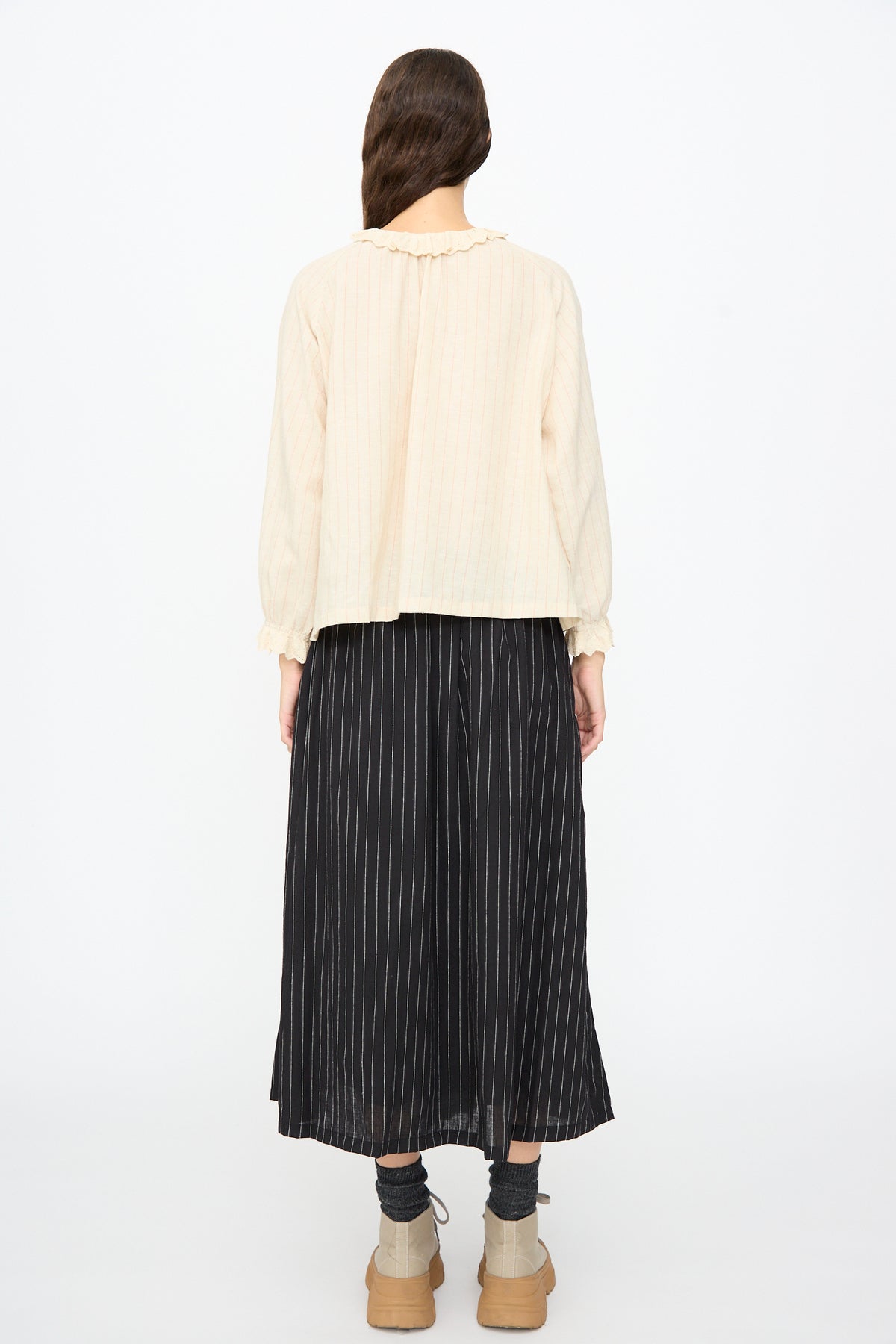 A person seen from behind wears an Ichi Antiquités Pinstripe Shirt in Natural paired with a black pinstriped midi skirt, dark socks, and light-colored boots.