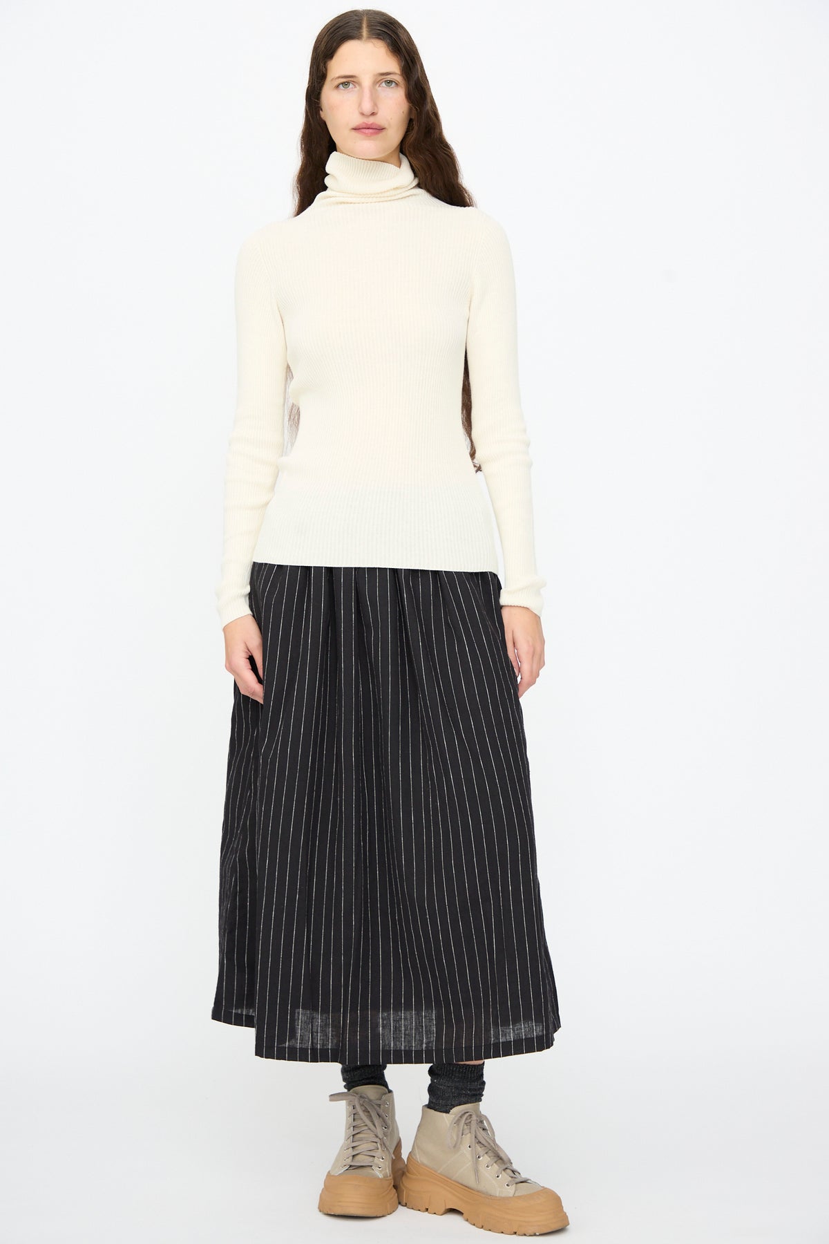 A person wearing a white turtleneck sweater, an Ichi Antiquités Pinstripe Skirt in Black, and beige lace-up boots.