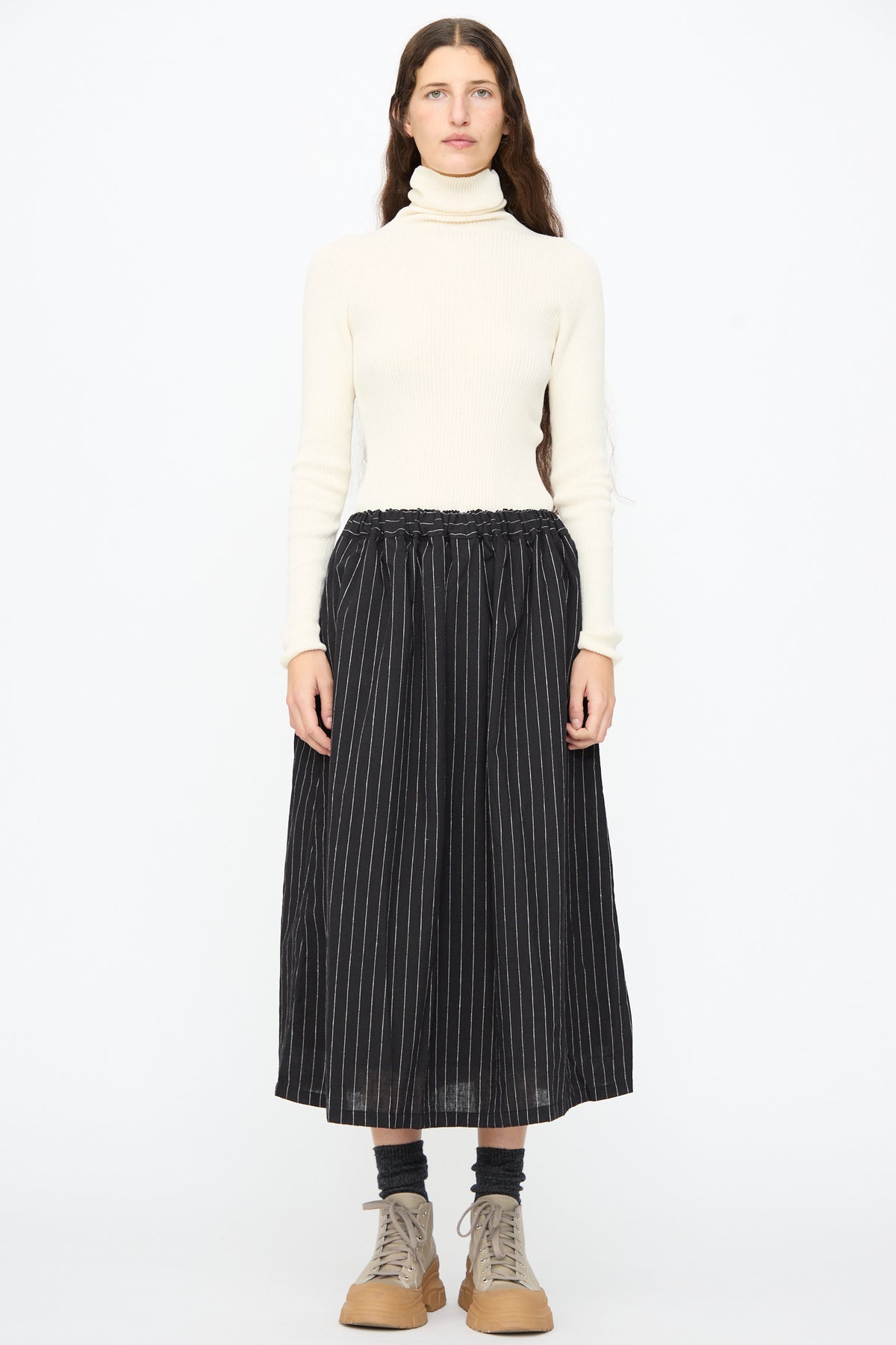 A person wearing a cream turtleneck sweater and an Ichi Antiquités Pinstripe Skirt in Black, featuring a long A-line midi silhouette with a gathered, elasticized waist.