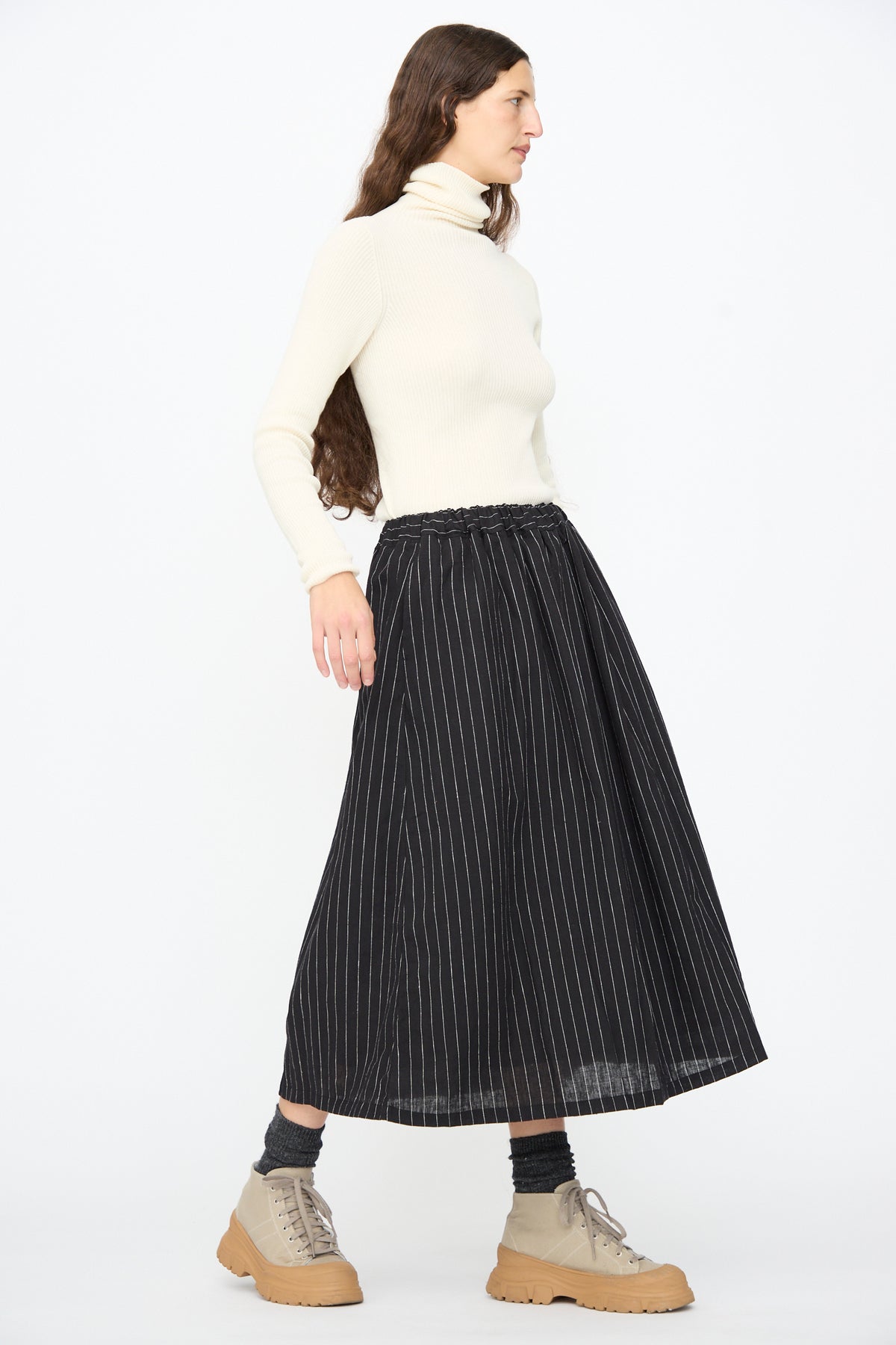 A person wears a cream turtleneck sweater, an Ichi Antiquités Pinstripe Skirt in Black, grey socks, and tan lace-up boots with thick rubber soles.