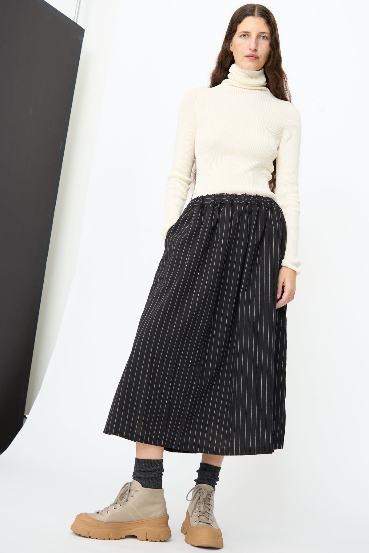 A person wearing a cream turtleneck sweater, an Ichi Antiquités Pinstripe Skirt in Black, and beige lace-up boots.