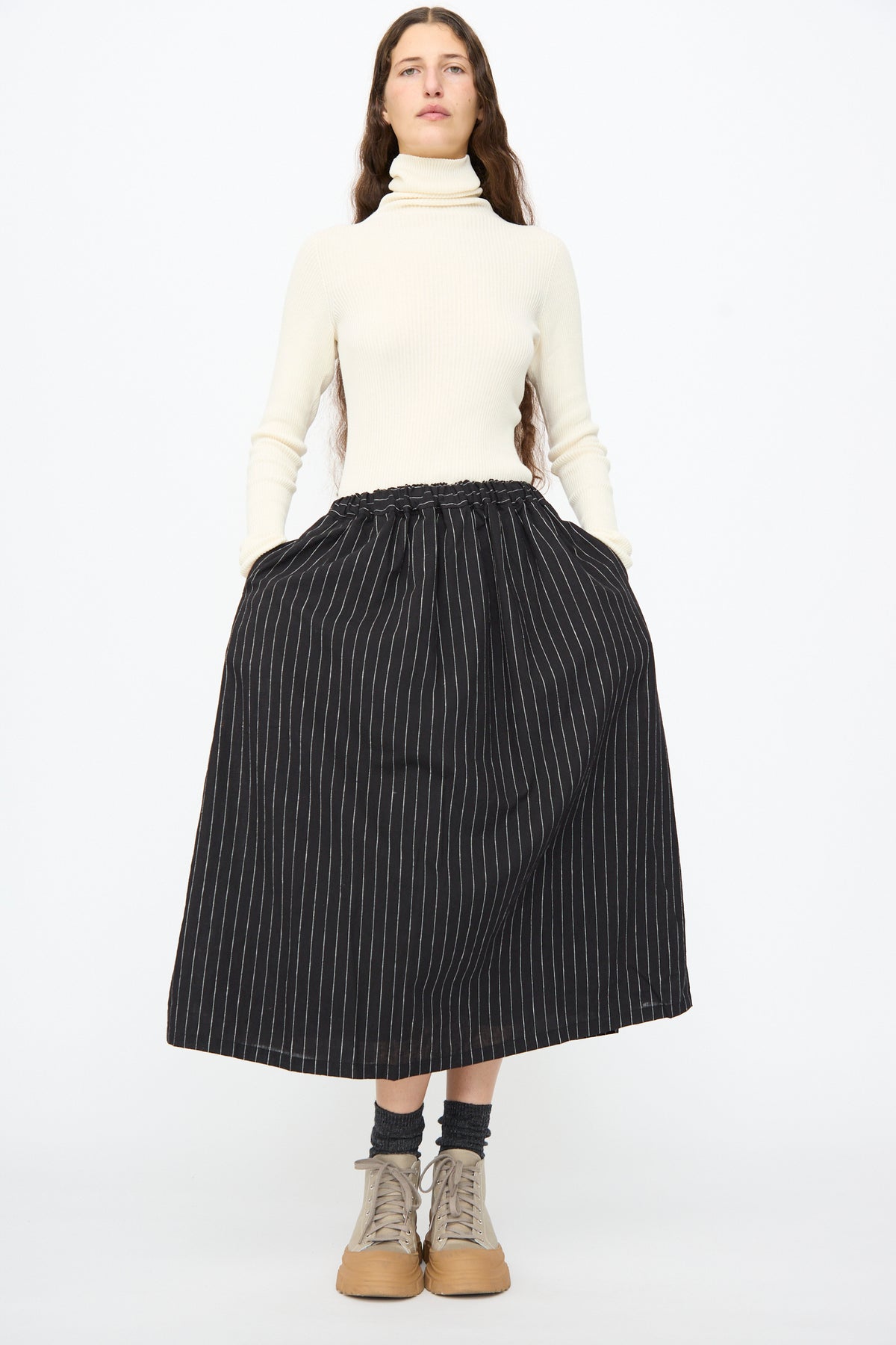 A person wearing a cream turtleneck sweater and an Ichi Antiquités Pinstripe Skirt in Black, paired with gray socks and beige sneakers.