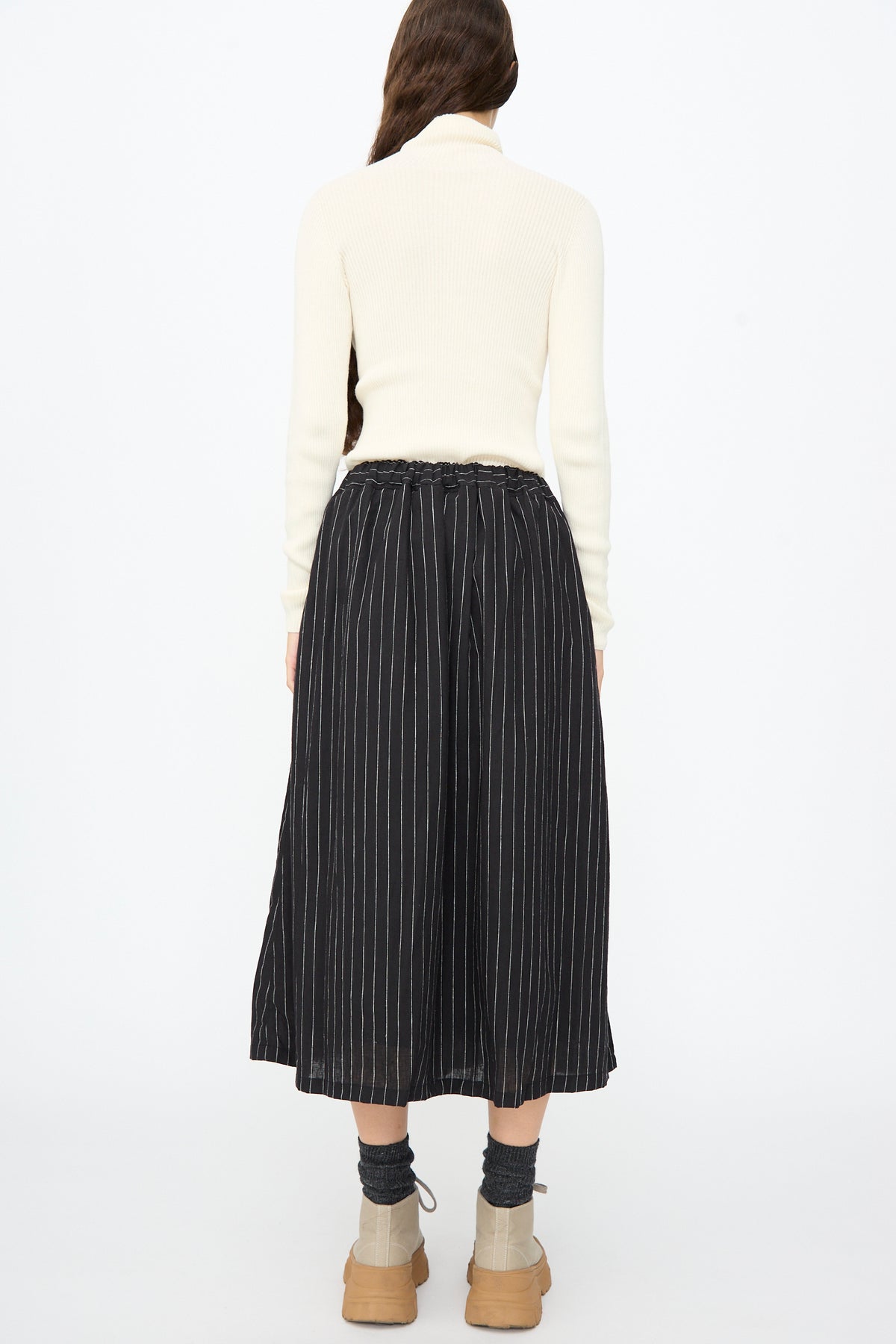 A person seen from behind wears a white turtleneck sweater and an Ichi Antiquités Pinstripe Skirt in Black, paired with dark socks and tan shoes.