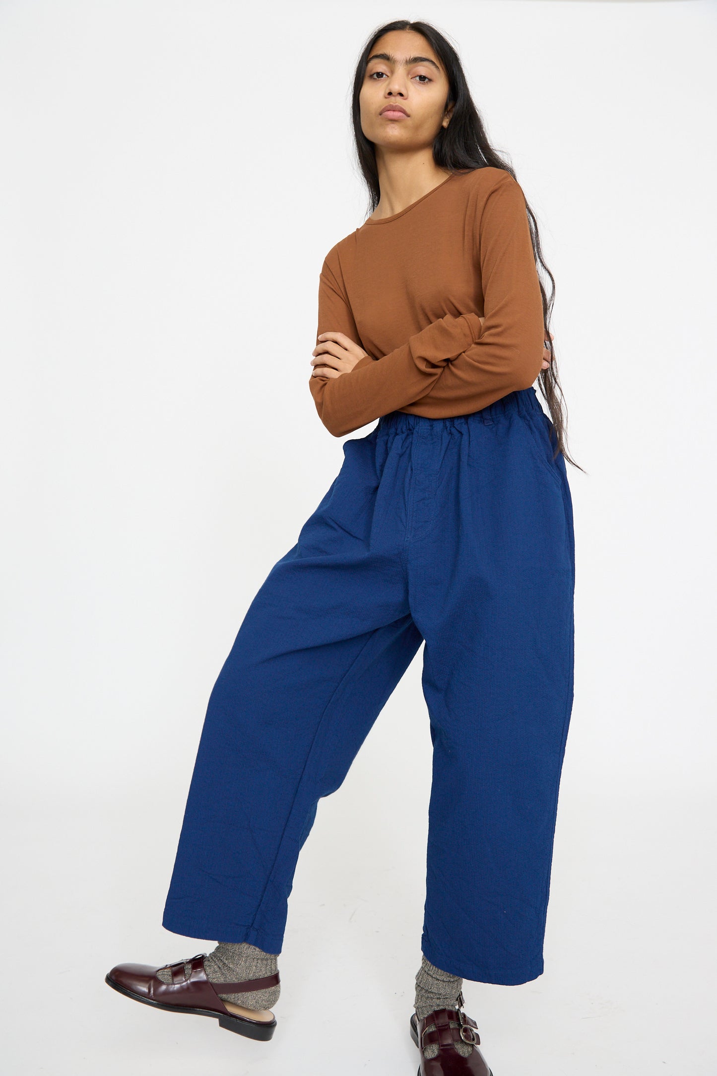 A person stands relaxed against a white background, dressed in a brown long-sleeve shirt, Sashiko Pants in Indigo by Ichi Antiquités, gray socks, and brown shoes.