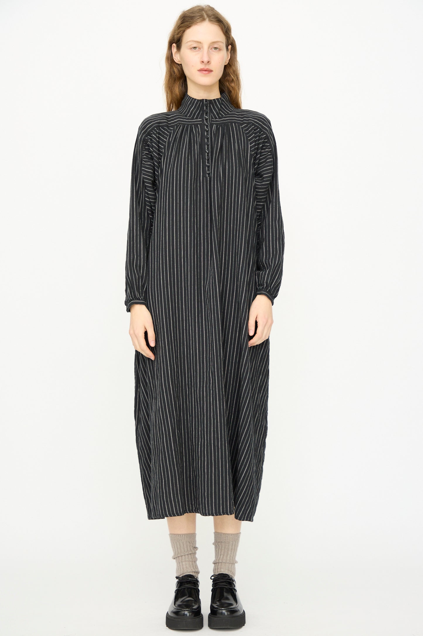 A person faces forward wearing the Ichi Antiquités Shirring Brushed Stripe Dress in Black, featuring white vertical stripes, paired with beige socks and black shoes against a plain white background.