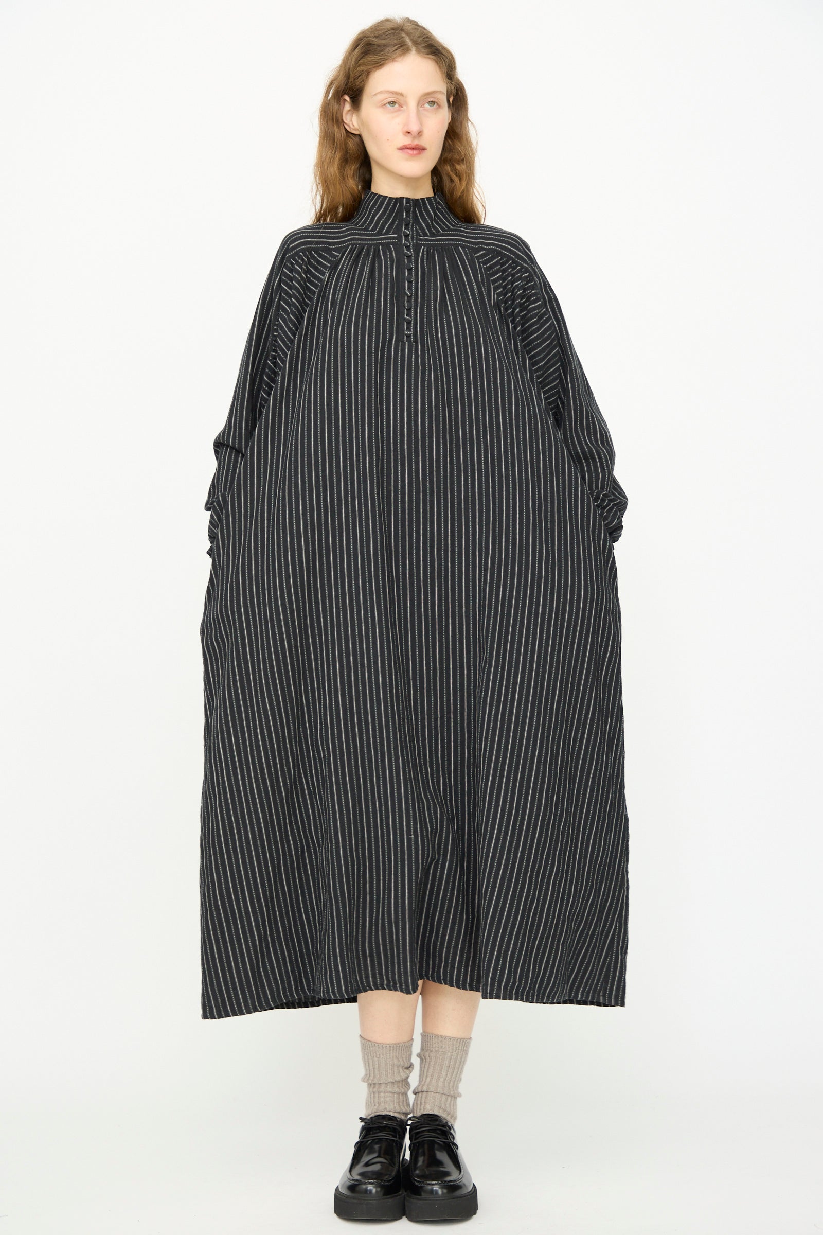 A person stands facing forward against a plain white background wearing the Ichi Antiquités Shirring Brushed Stripe Dress in Black, a loose-fitting, long cotton maxi dress with white pinstripes. Made in Japan.