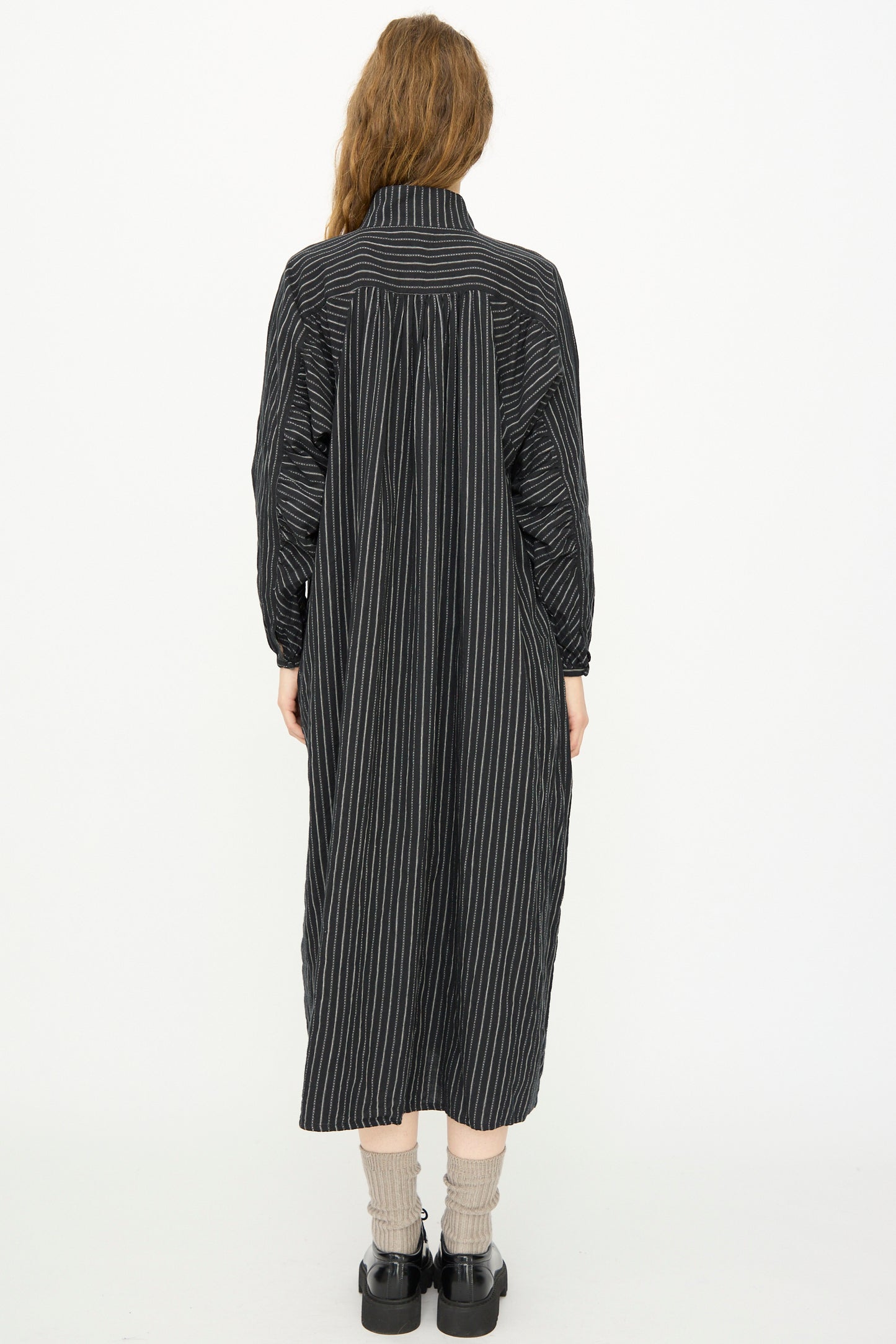 A person with long hair wears the Ichi Antiquités Shirring Brushed Stripe Dress in Black, paired with gray socks and black shoes, standing with their back to the camera against a white background.