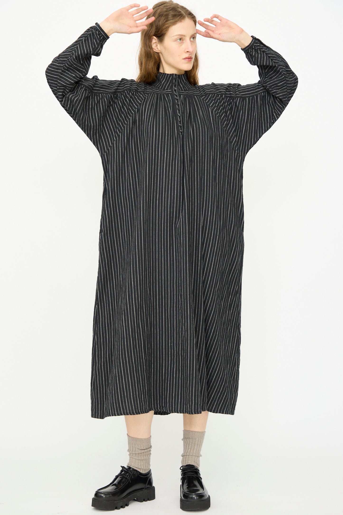 A person stands with arms raised, wearing the Ichi Antiquités Shirring Brushed Stripe Dress in Black, paired with beige socks and black shoes, against a plain white background.