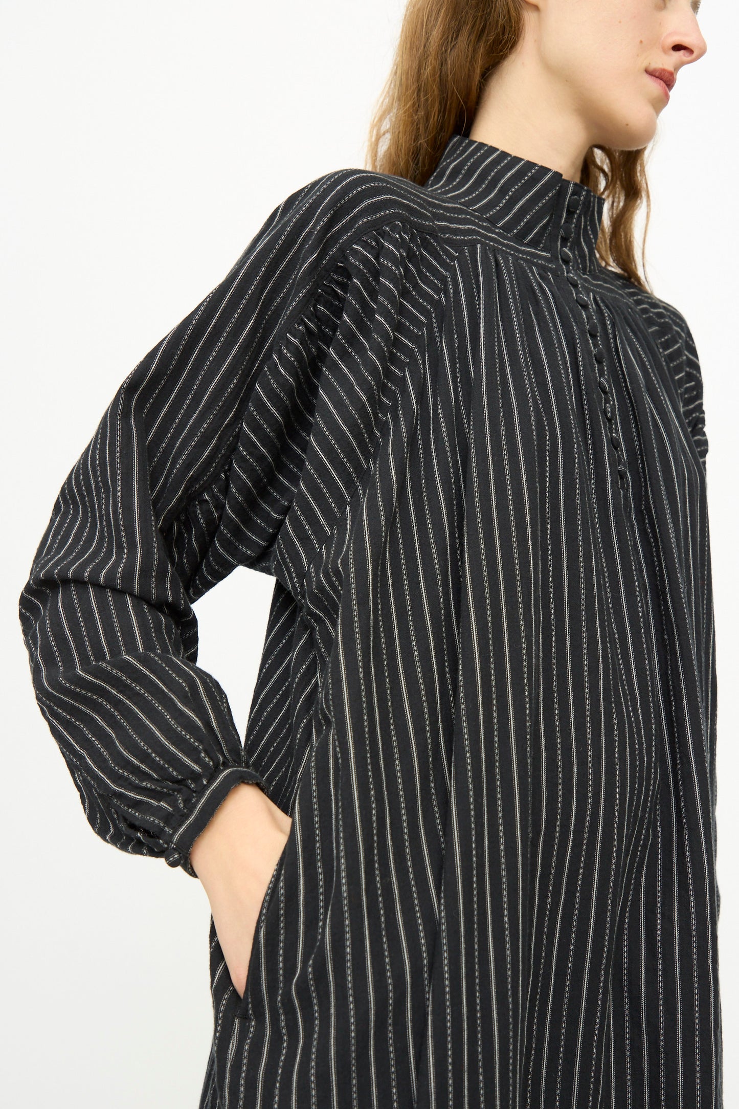 A person wearing the Shirring Brushed Stripe Dress in Black by Ichi Antiquités, a long-sleeved maxi dress with white vertical pinstripes and a high collar, stands with one hand in their pocket.