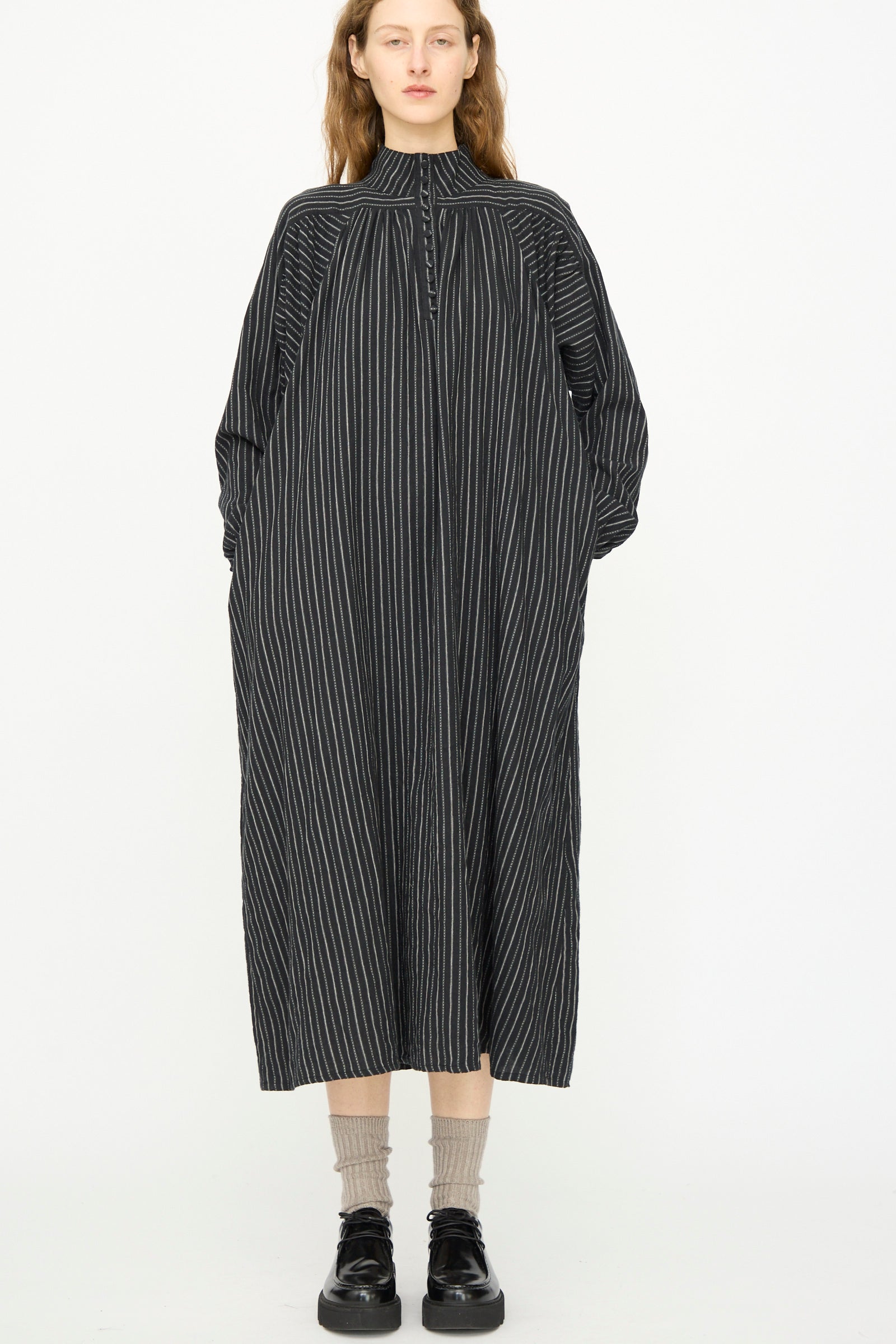 A person faces forward wearing the Ichi Antiquités Shirring Brushed Stripe Dress in Black with beige socks and black shoes, posed against a plain white background.