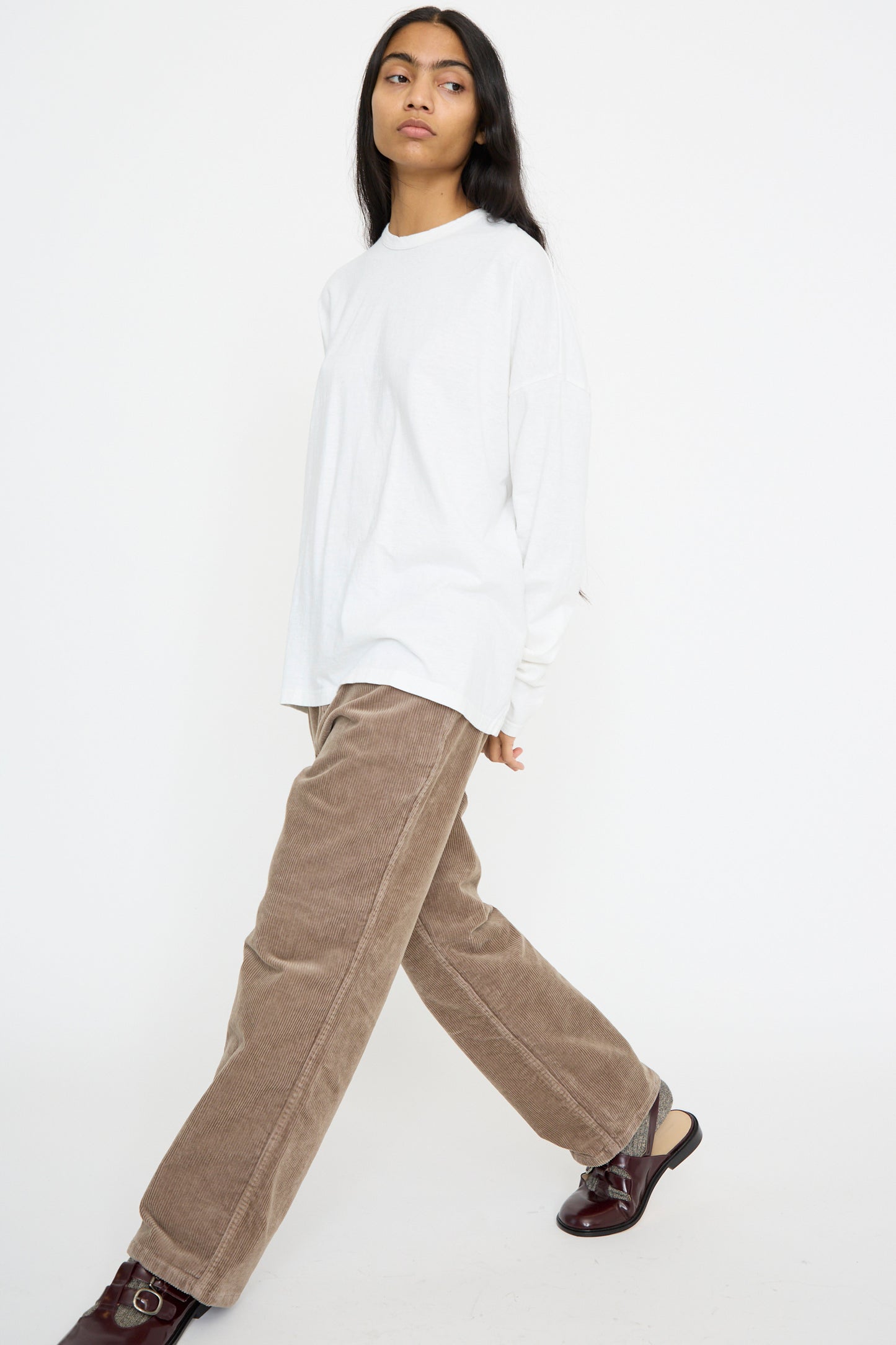 Person in a white long-sleeve shirt, Ichi Antiquités' Stretch Call Corduroy Pants in Mocha, and brown shoes, walks against a plain white backdrop.