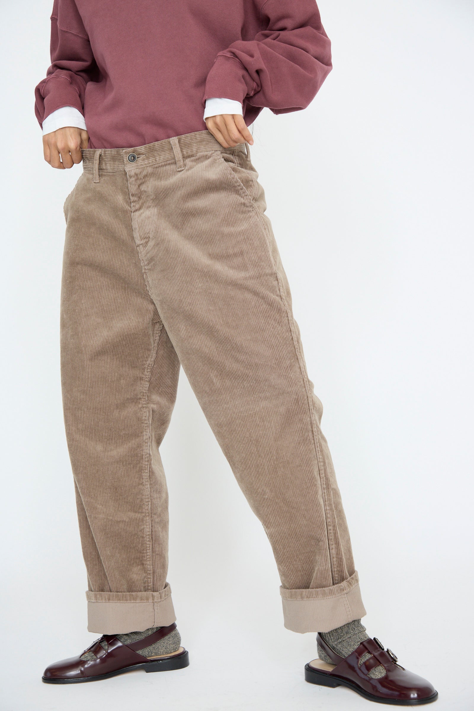 A person wears a burgundy sweater and relaxed fit, mid-waist Stretch Call Corduroy Pants in Mocha by Ichi Antiquités. Paired with gray socks and burgundy loafers, set against a white background and cropped from the bust down.