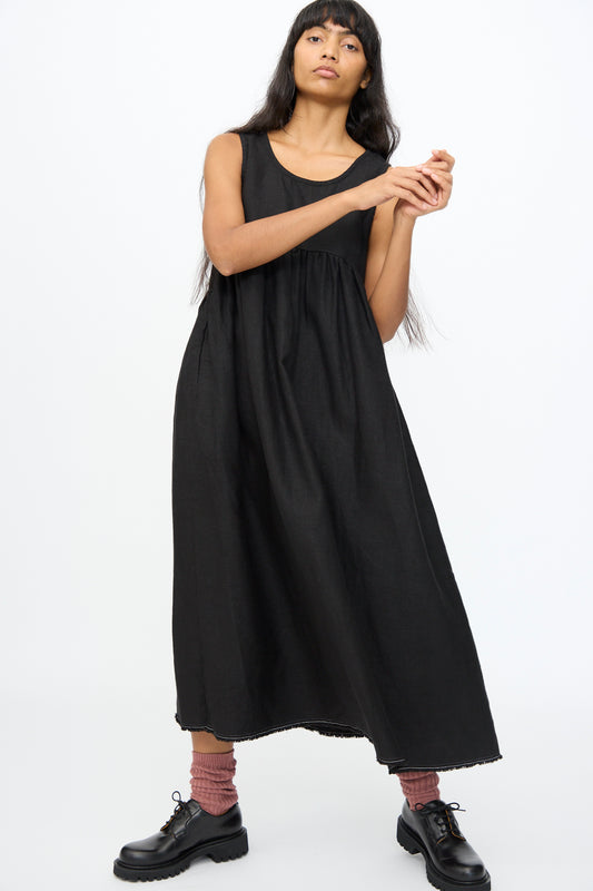 A woman wearing the Ichi Antiquités Twill Weave Linen Dress in Black stands against a plain white background, paired with black shoes and pink socks.