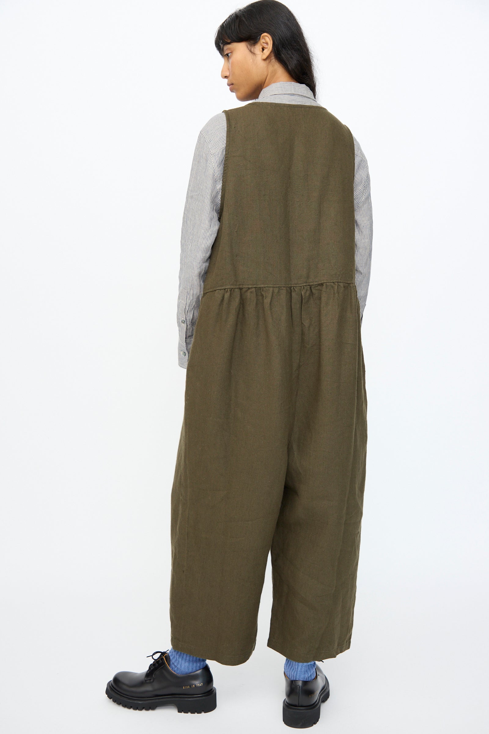 A person stands with their back to the camera, wearing the Ichi Antiquités Twill Weave Linen Jumpsuit in Khaki over a gray long-sleeve shirt, blue socks, and black shoes, set against a plain white background.