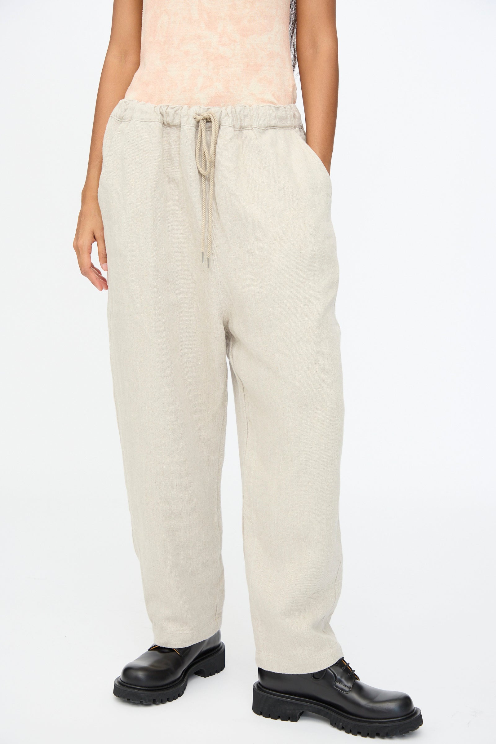 A person wears Ichi Antiquités Twill Weave Linen Pant in Natural with a drawstring waist and black lace-up shoes, hands in pockets, standing against a plain white background.