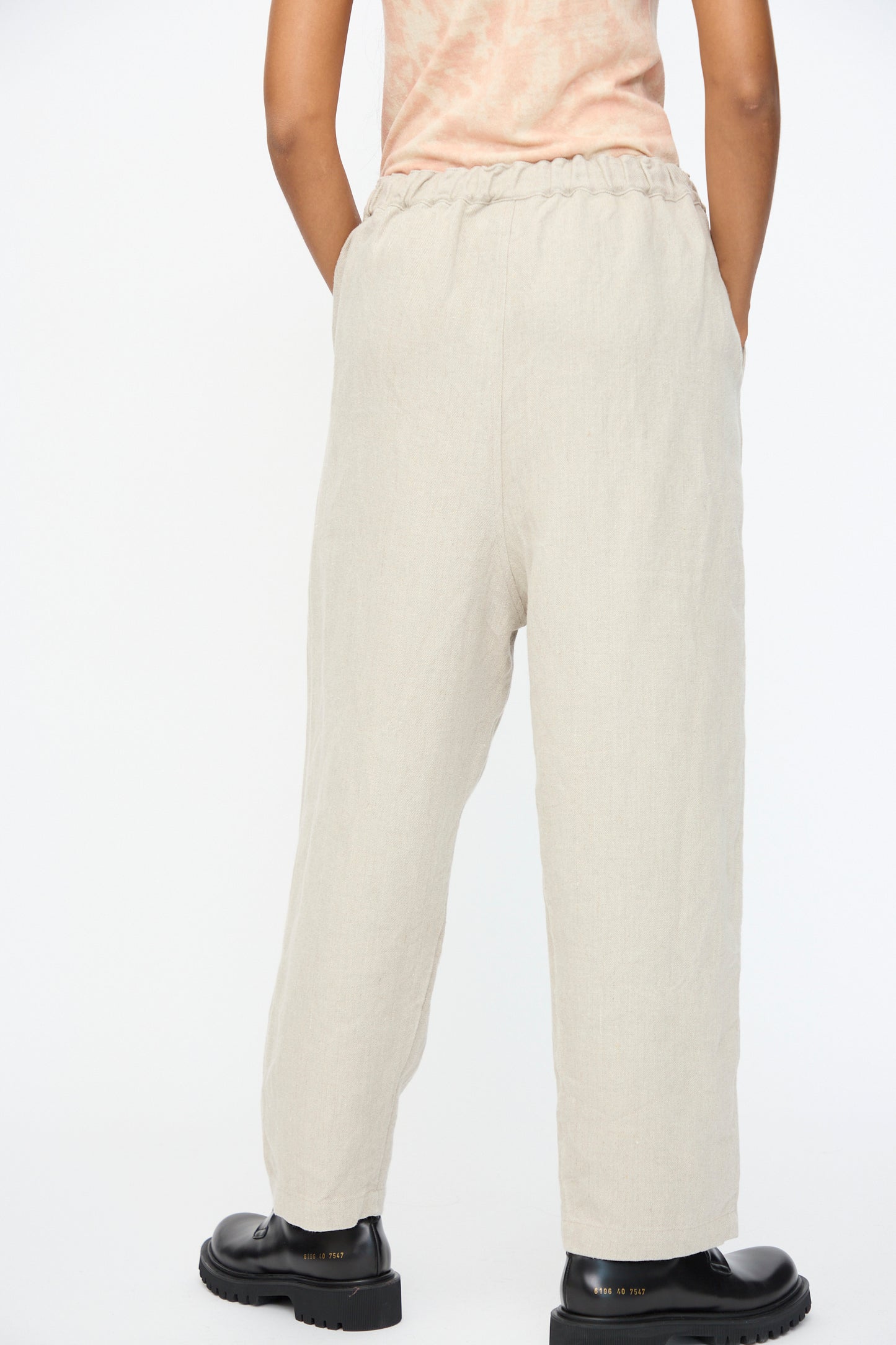 A person wearing Ichi Antiquités Twill Weave Linen Pant in Natural, featuring a loose wide-leg fit and drawstring waistband, with black shoes and hands in pockets, photographed from behind against a plain white background.