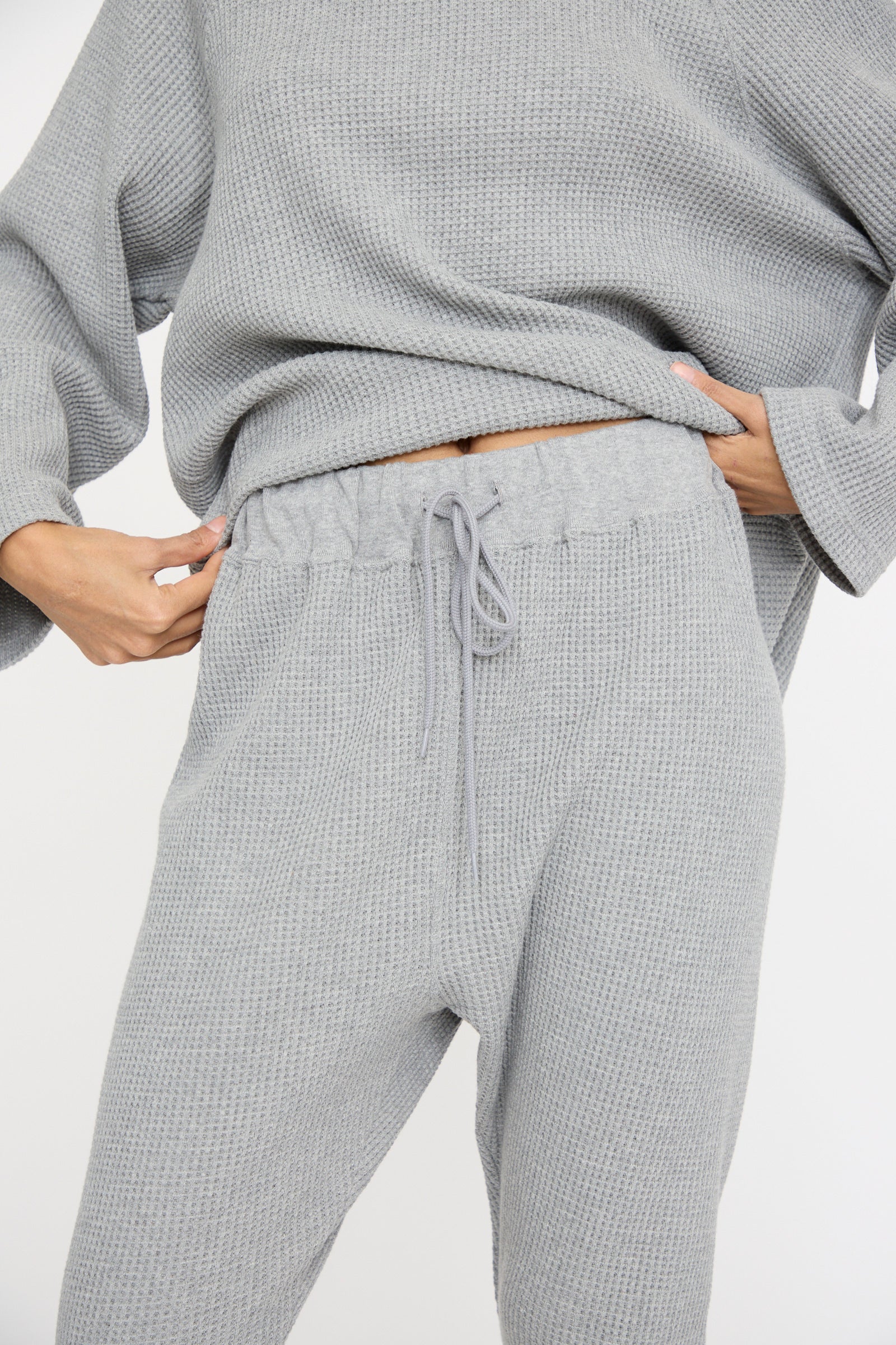 A close up of a person wearing Ichi Antiquités Waffle Knit Pants in Gray, holding the waistband of the slim fit loungewear set.