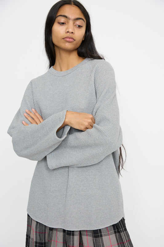 A person with long hair stands with arms crossed against a light background, wearing an Ichi Antiquités Waffle Knit Pullover in Gray and a plaid skirt. 