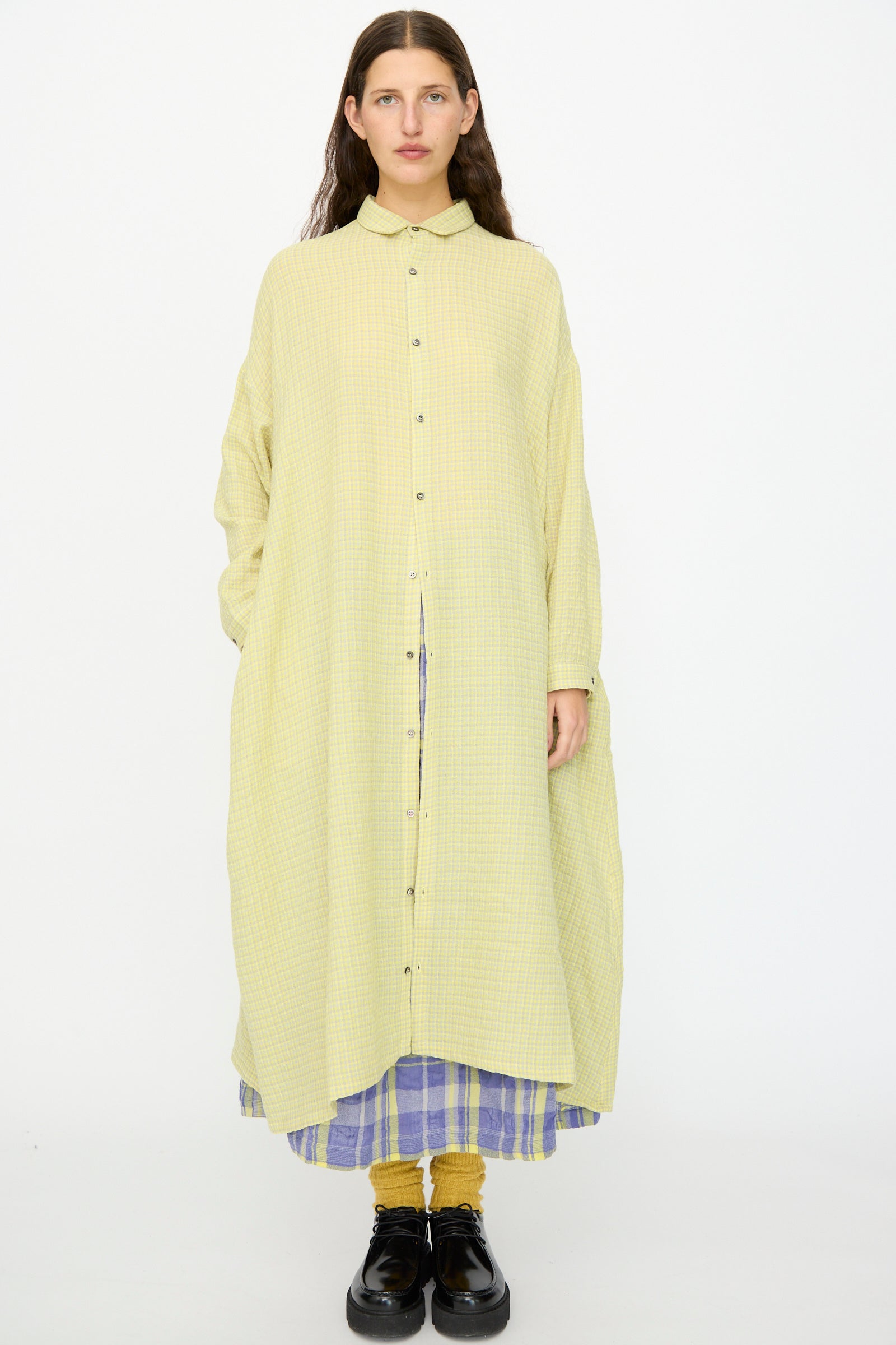 A person stands against a plain background wearing the Ichi Antiquités Wool Cotton Gingham Dress in Yellow and Gray, styled with a purple checkered skirt, yellow socks, and black shoes.
