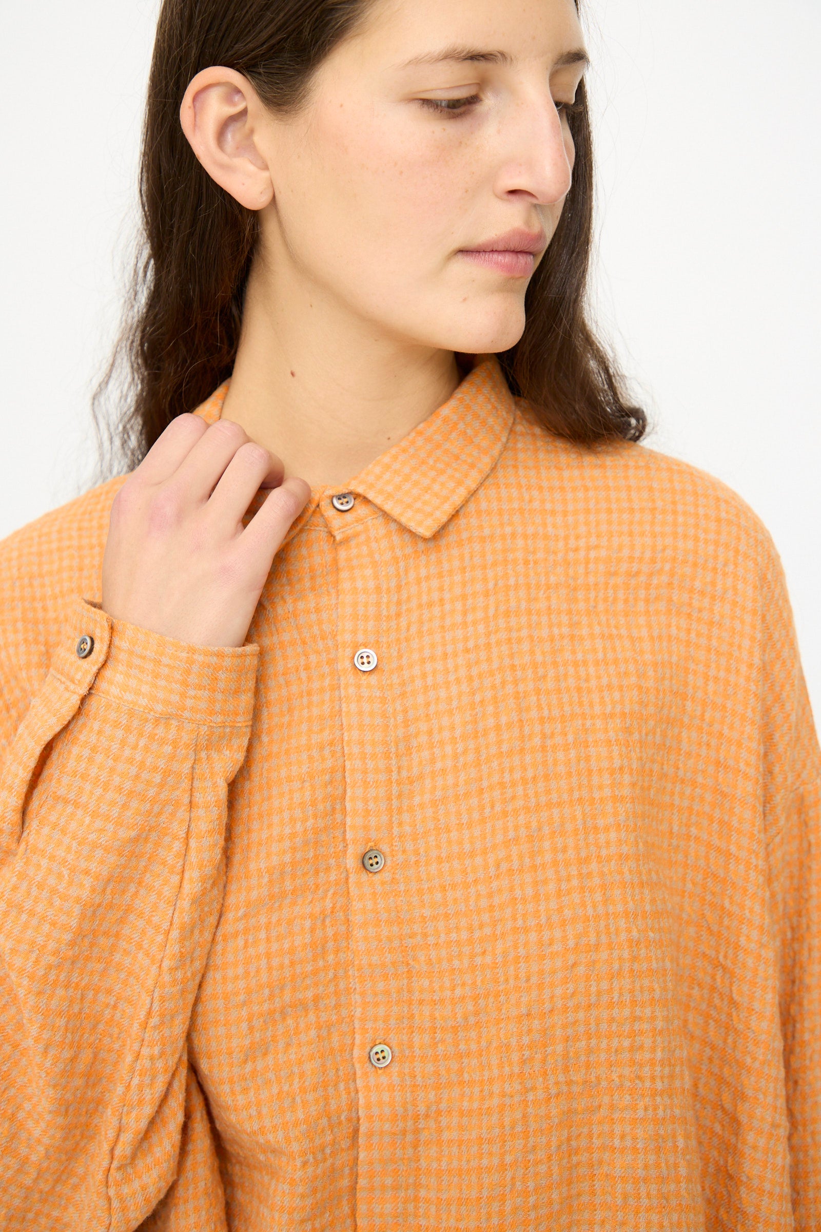 A person with long brown hair wears the Ichi Antiquités Wool Cotton Gingham Shirt in Orange and Mocha, made in Japan, looking down and to the side while holding the collar with one hand.