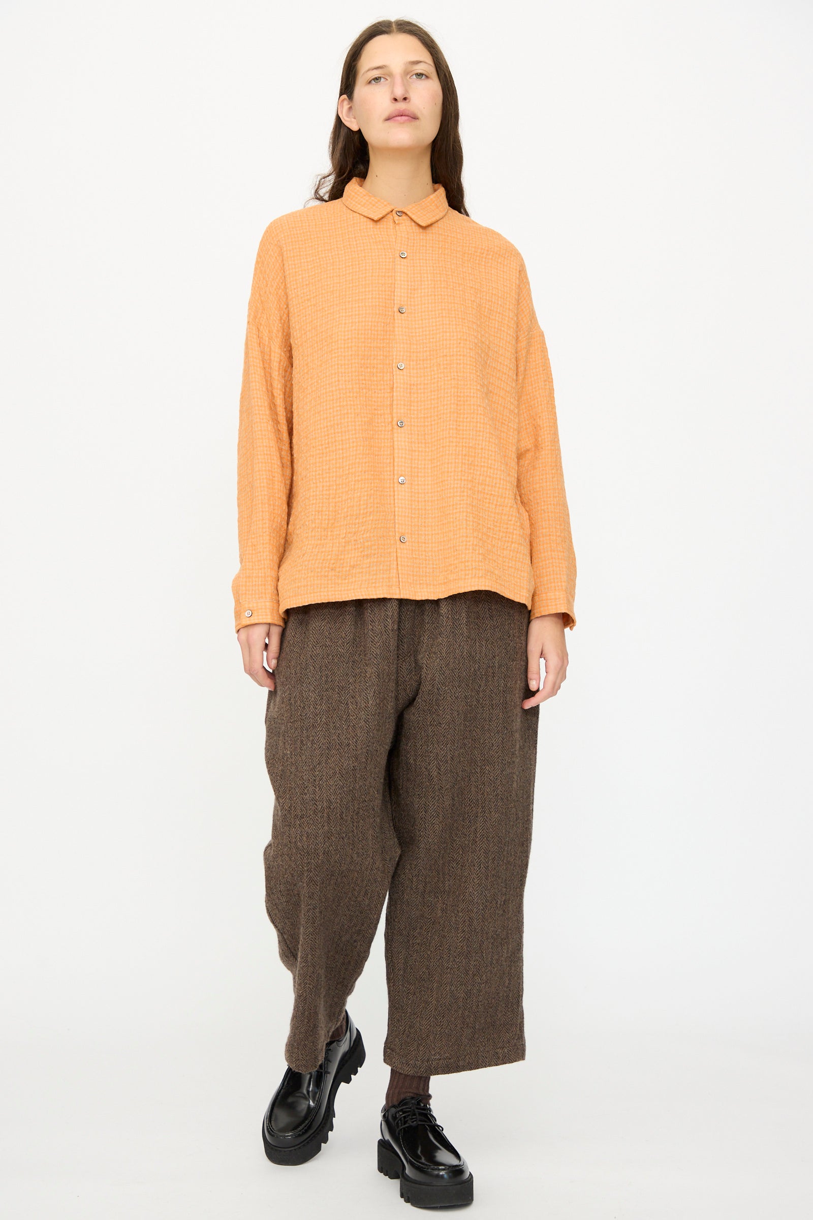 A person stands against a plain background wearing the Ichi Antiquités Wool Cotton Gingham Shirt in Orange and Mocha, paired with loose brown pants and black platform shoes.
