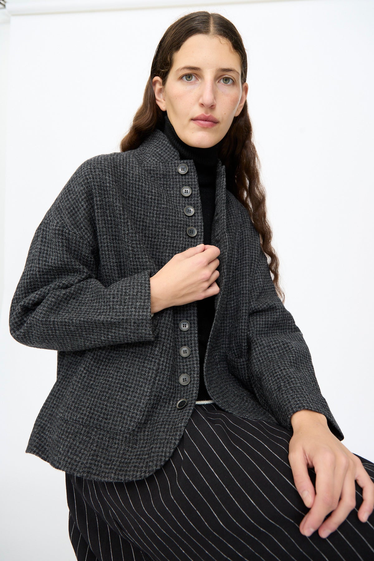 A person wears an Ichi Antiquités Wool Gauze Houndstooth Jacket in Charcoal over a black turtleneck and black pinstriped trousers.