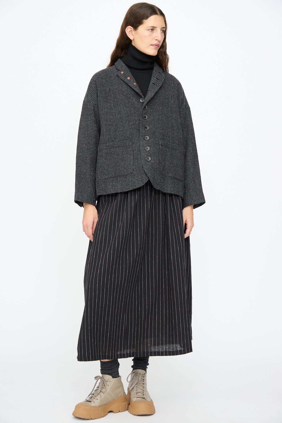 A person wearing an Ichi Antiquités Wool Gauze Houndstooth Jacket in Charcoal over a black turtleneck and a black pinstriped maxi skirt, with grey socks and tan lug-sole boots.