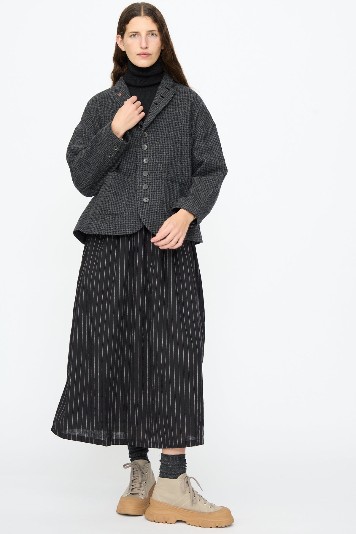 A person wears a relaxed fit, charcoal Wool Gauze Houndstooth Jacket in Charcoal by Ichi Antiquités over a black turtleneck, a long black pinstripe skirt, and tan lace-up sneakers.