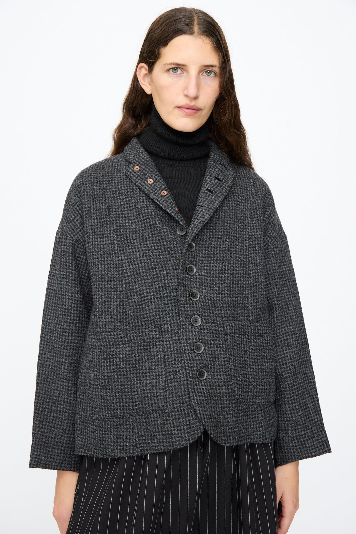 A person wears an Ichi Antiquités Wool Gauze Houndstooth Jacket in Charcoal, featuring a notched collar and front buttons, over a black turtleneck and pinstriped trousers.