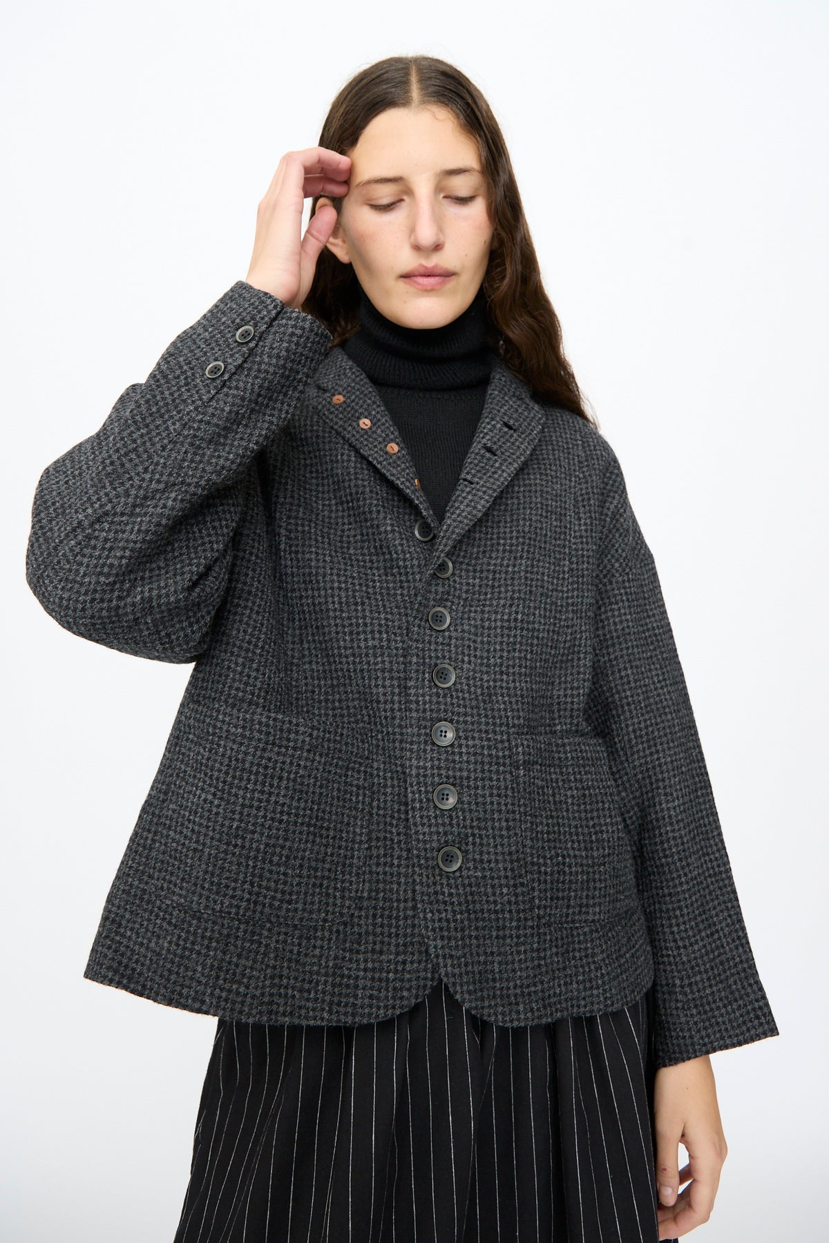 A person wears an Ichi Antiquités Wool Gauze Houndstooth Jacket in Charcoal, a charcoal grey houndstooth wool jacket in a relaxed, single-breasted fit layered over a black turtleneck.