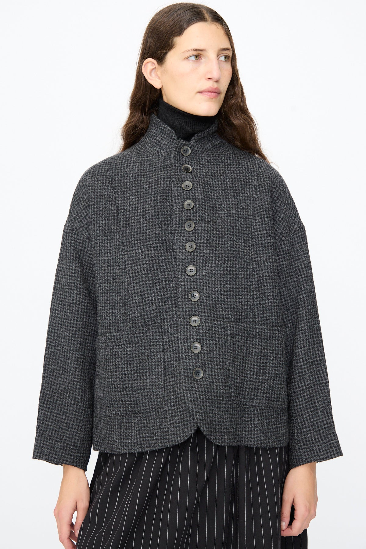 A person wearing an Ichi Antiquités Wool Gauze Houndstooth Jacket in Charcoal, a relaxed-fit wool jacket with a high button-up collar and long sleeves.